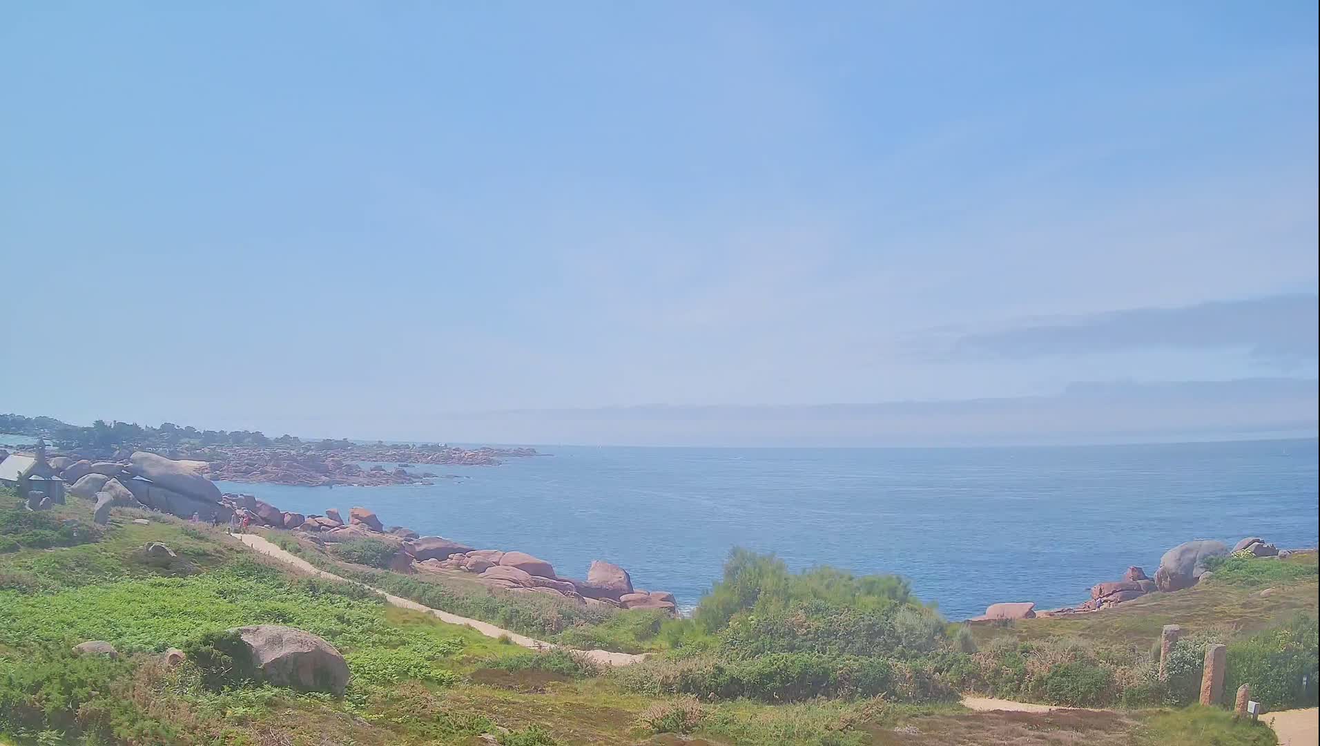 A coastal path winds through grassy hills dotted with large pink rocks overlooking a calm blue ocean under a clear, light-blue sky.