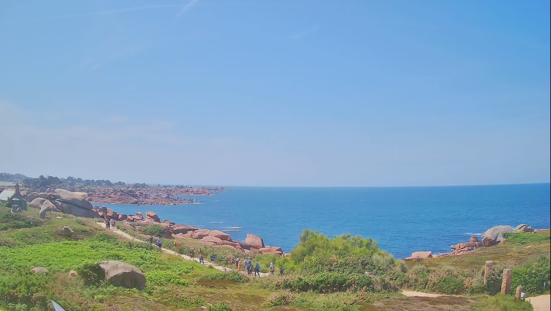 A coastal path winds through a rocky, grassy landscape overlooking a calm blue sea under a clear sky.