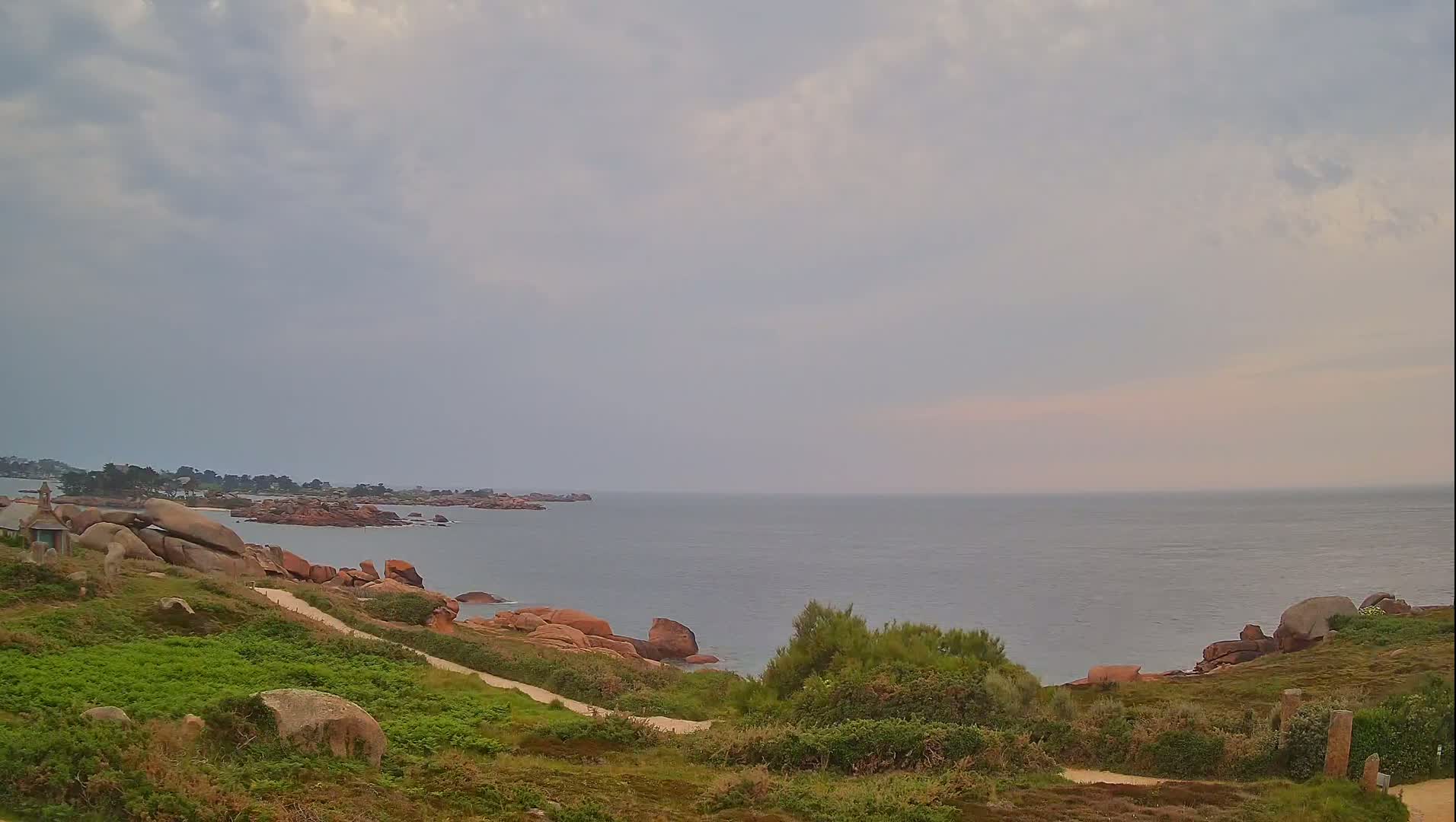 A coastal scene under a cloudy sky shows a path winding down a grassy hillside toward a rocky shoreline and calm sea.