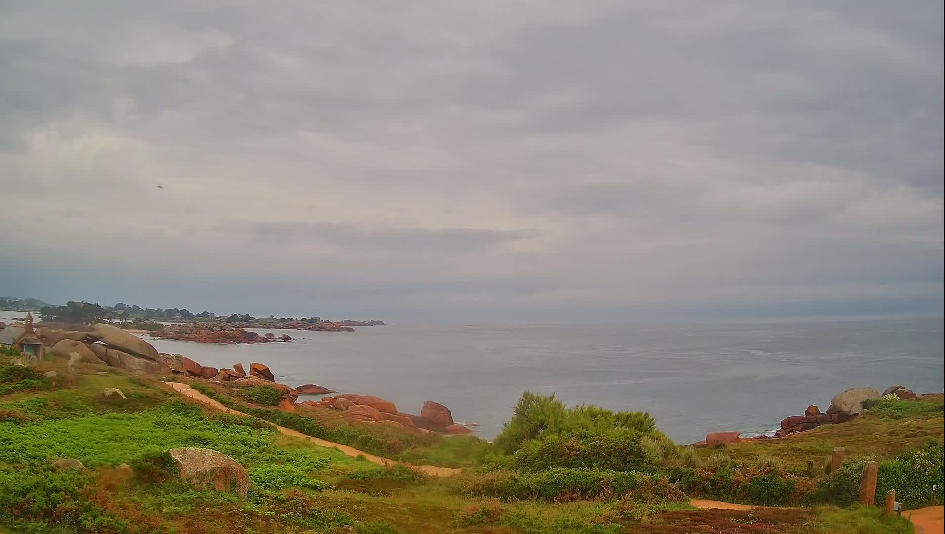 A coastal path winds through green vegetation alongside a rocky shoreline, overlooking a calm, gray sea under an overcast sky.
