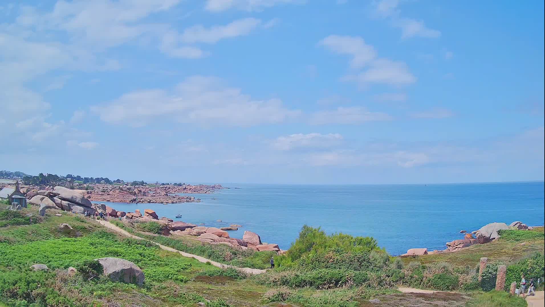 A partly cloudy sky overlooks a rocky coastline with a calm, blue ocean; a path winds through green vegetation near pink granite rocks where people are walking.