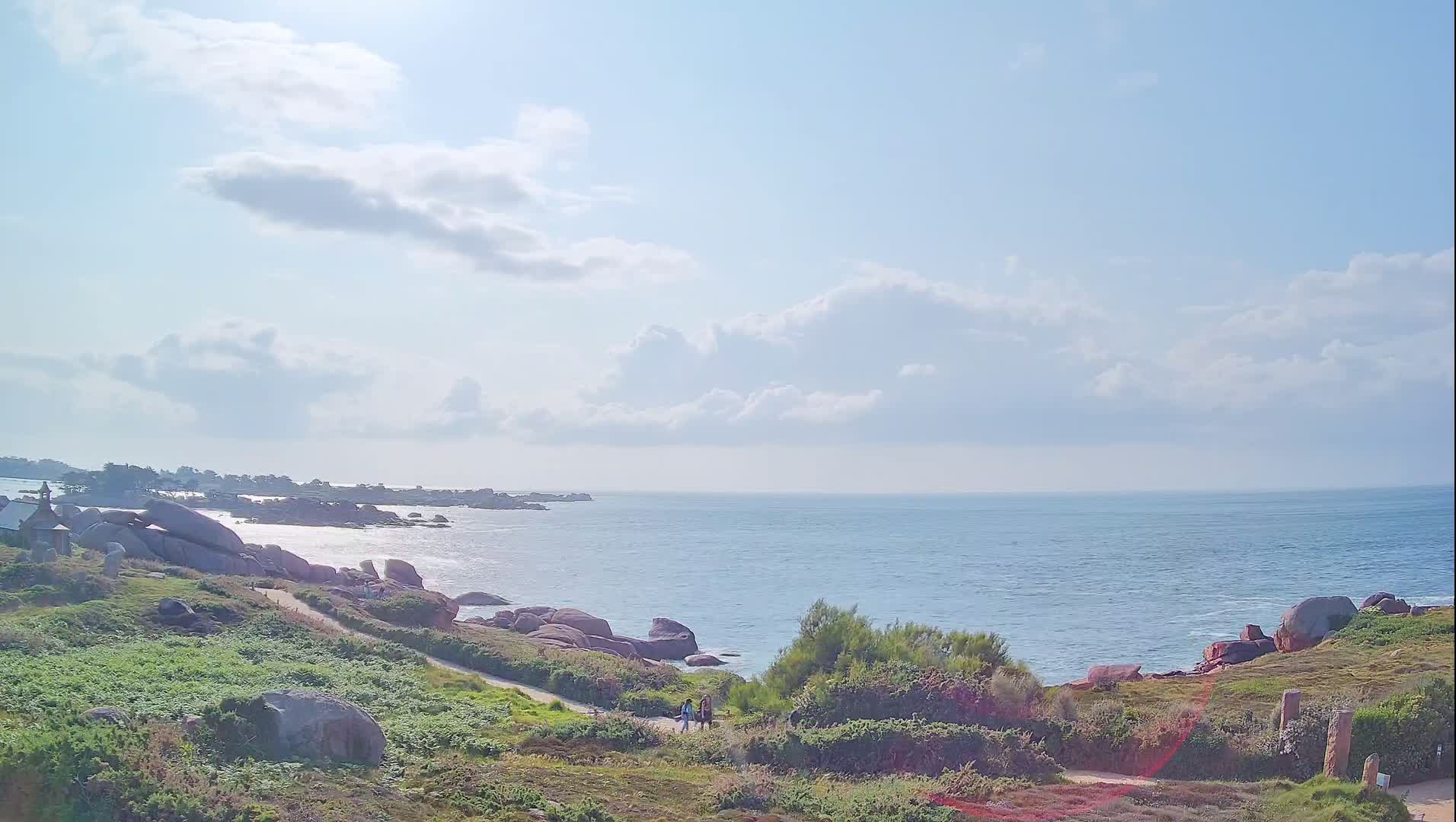 A partly cloudy day shows a coastal path winding through green vegetation toward a rocky shoreline and calm blue ocean.