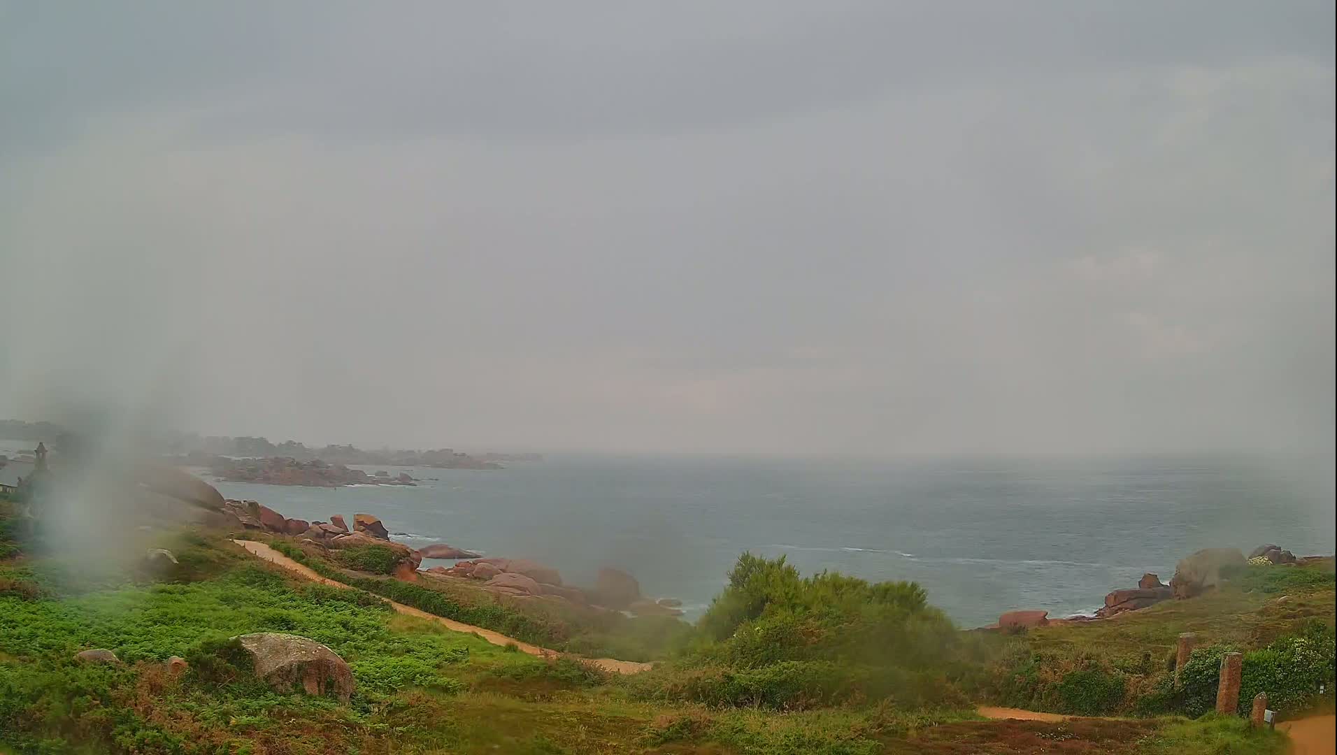 A rain-swept coastal scene shows a path winding down a grassy hillside toward a rocky shoreline and a hazy ocean under a gray sky.