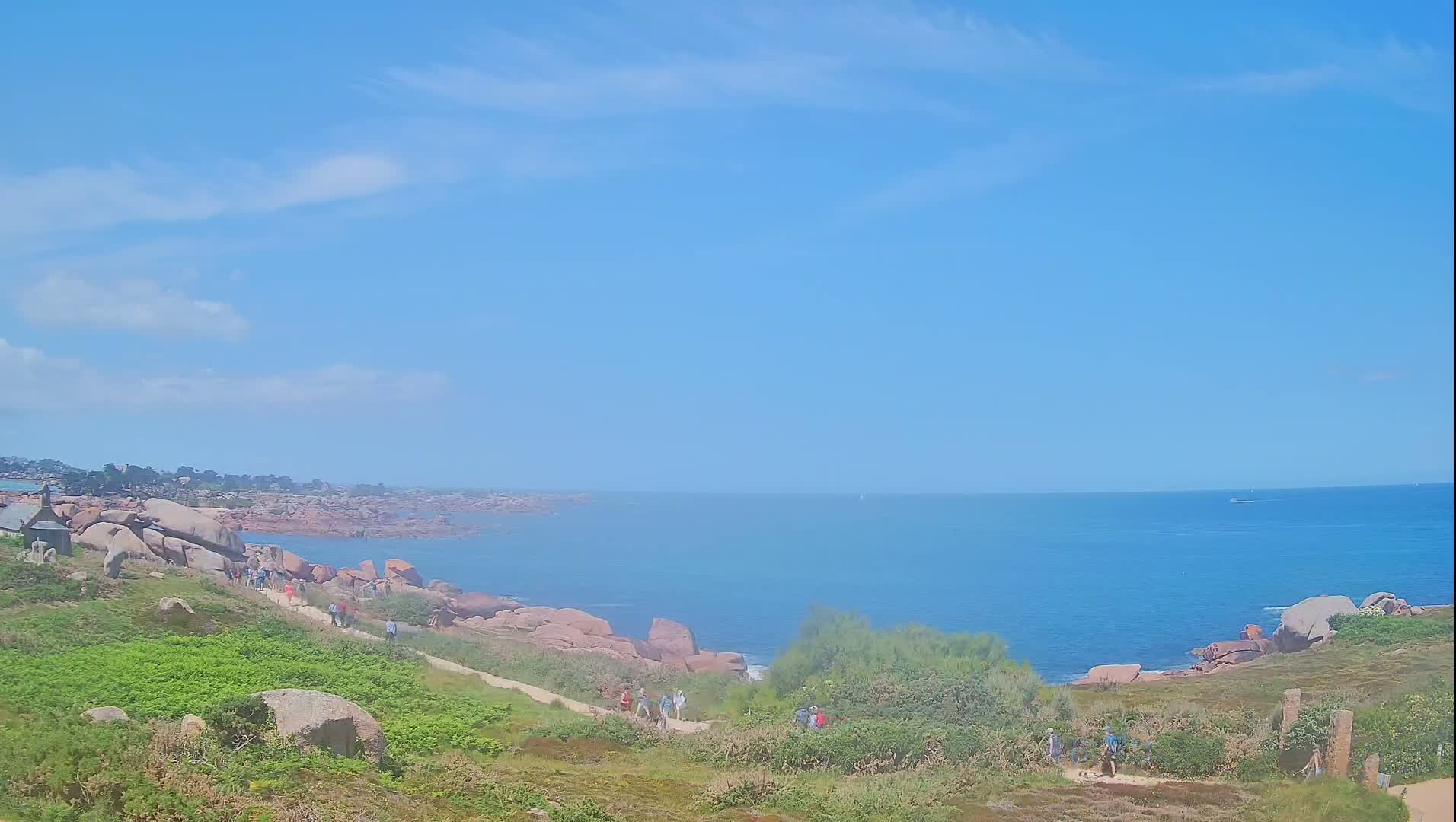 A coastal path winds through green vegetation and pink rocks toward a calm blue ocean under a mostly clear sky.