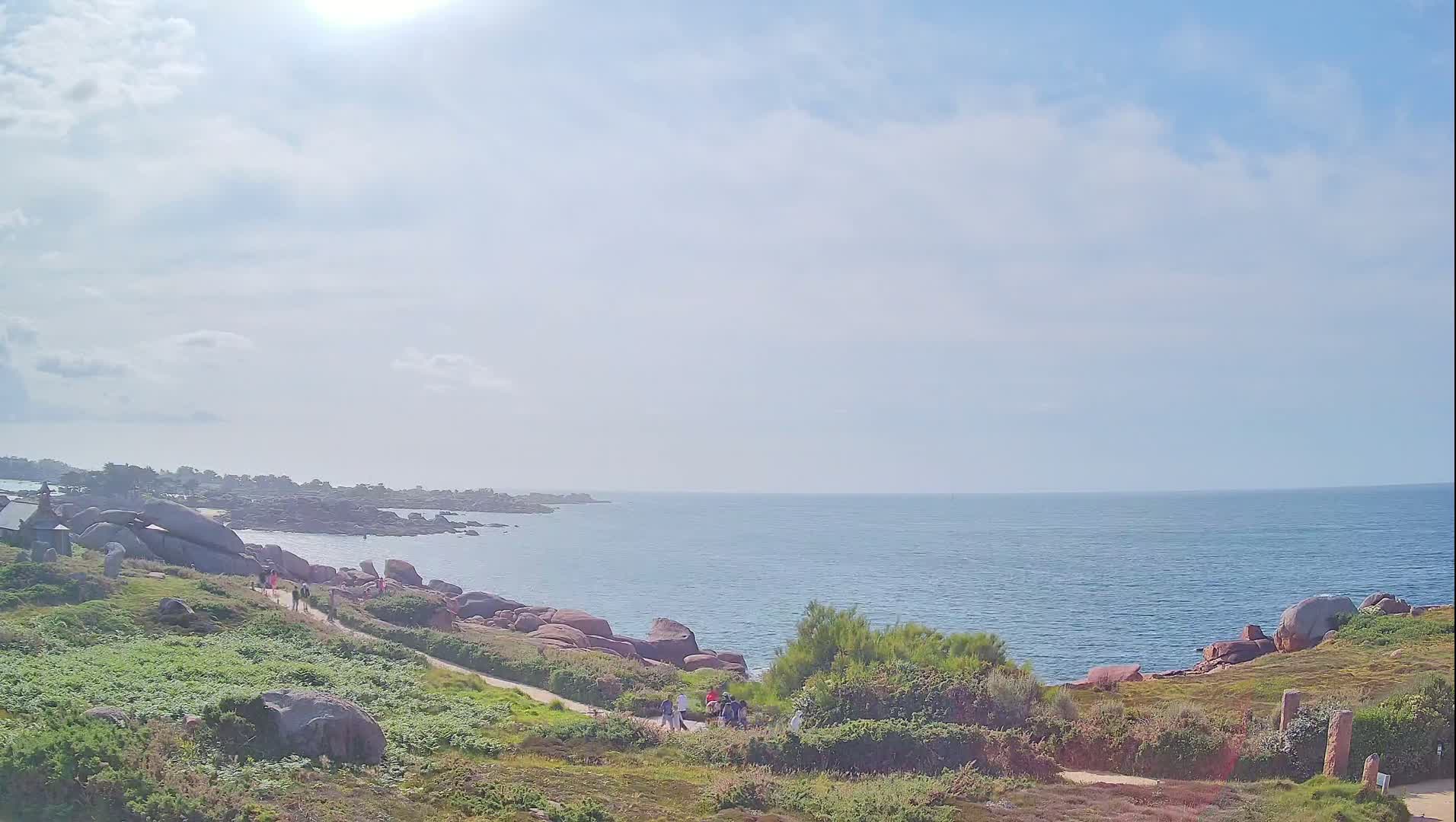 A sunny day reveals a coastal path winding through grassy terrain alongside a rocky shoreline and a calm blue ocean.