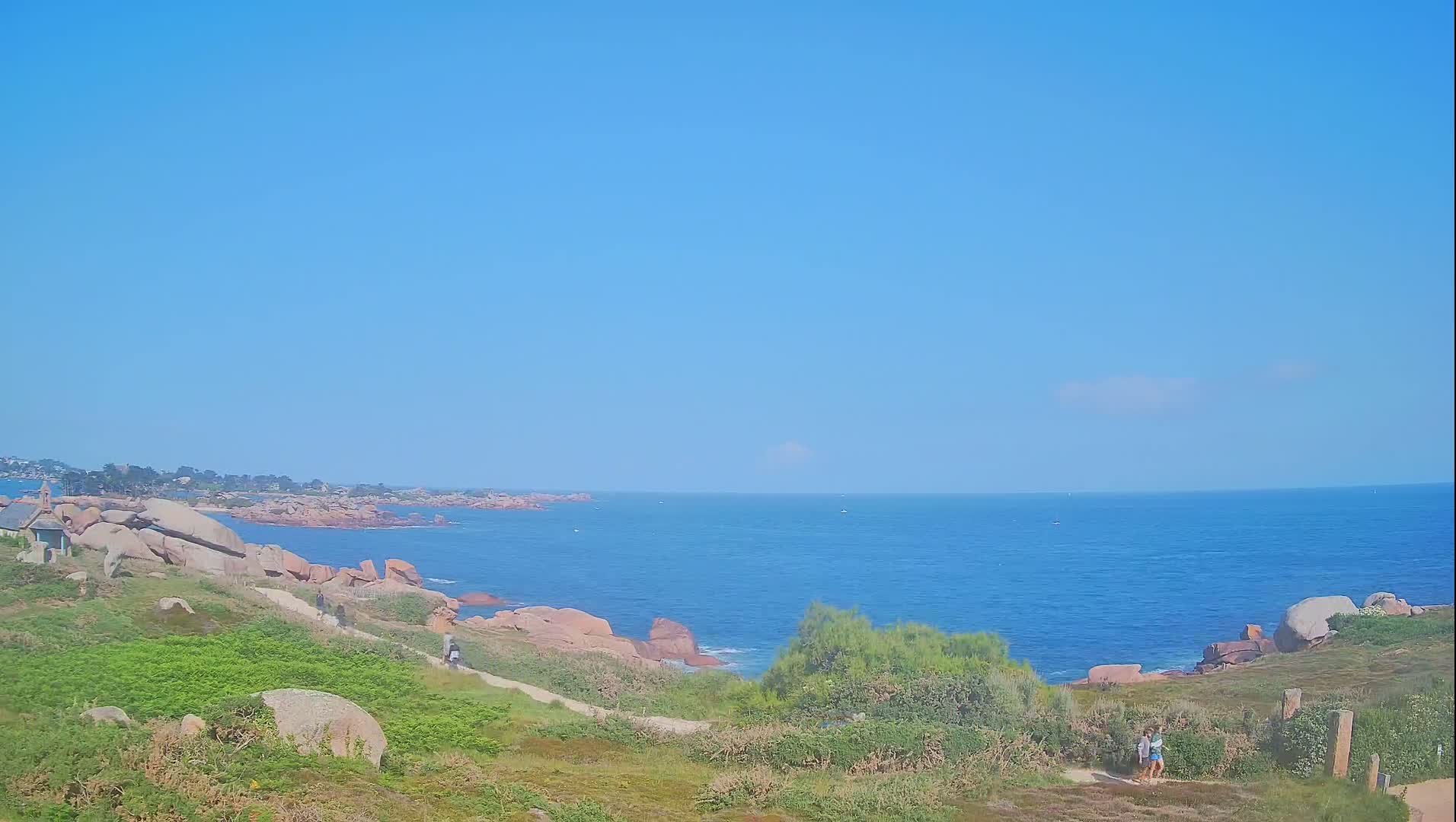 A sunny day reveals a coastal scene with pink granite rocks, a path winding through green vegetation towards a calm blue ocean.