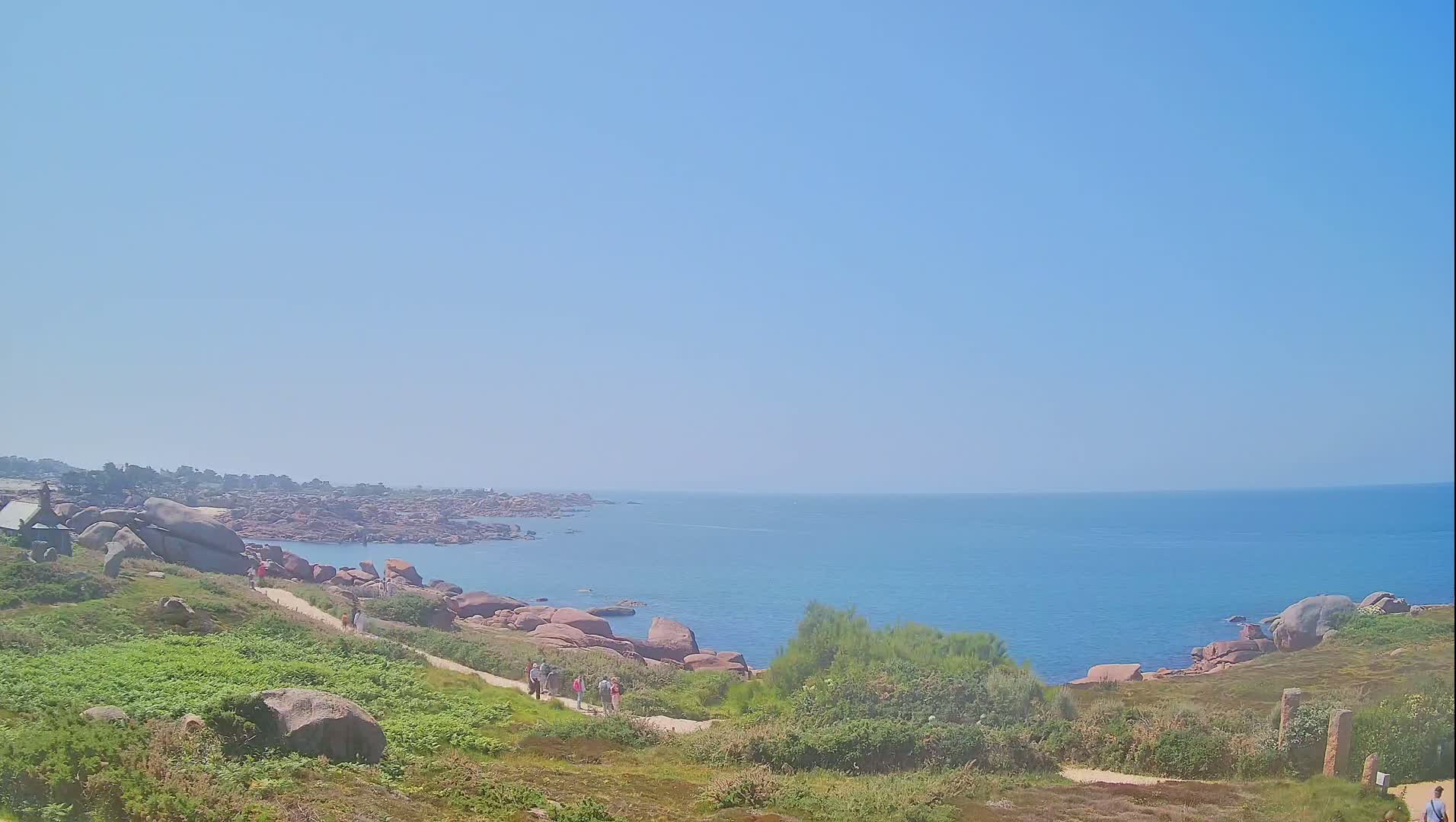 A coastal path winds through low-lying vegetation and pink granite rocks toward a calm, blue ocean under a clear sky.