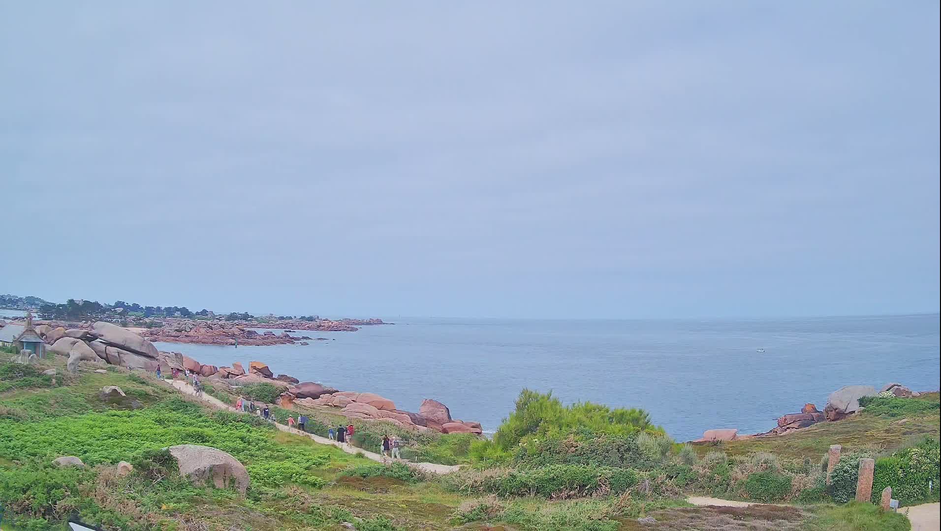 A coastal path winds through grassy terrain alongside pink rocks and a calm, light-blue ocean under an overcast sky.