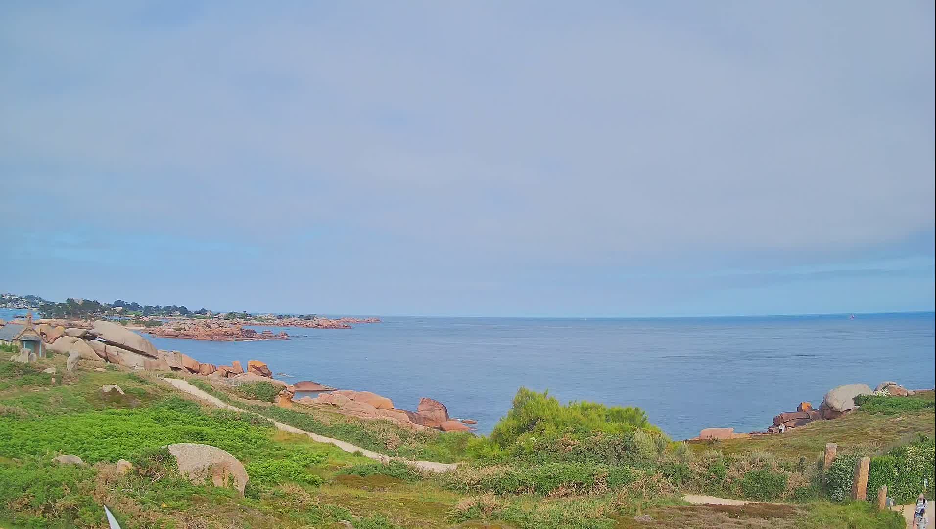 A tranquil coastal scene under a lightly clouded sky shows a calm sea meeting a rocky shoreline with green vegetation.