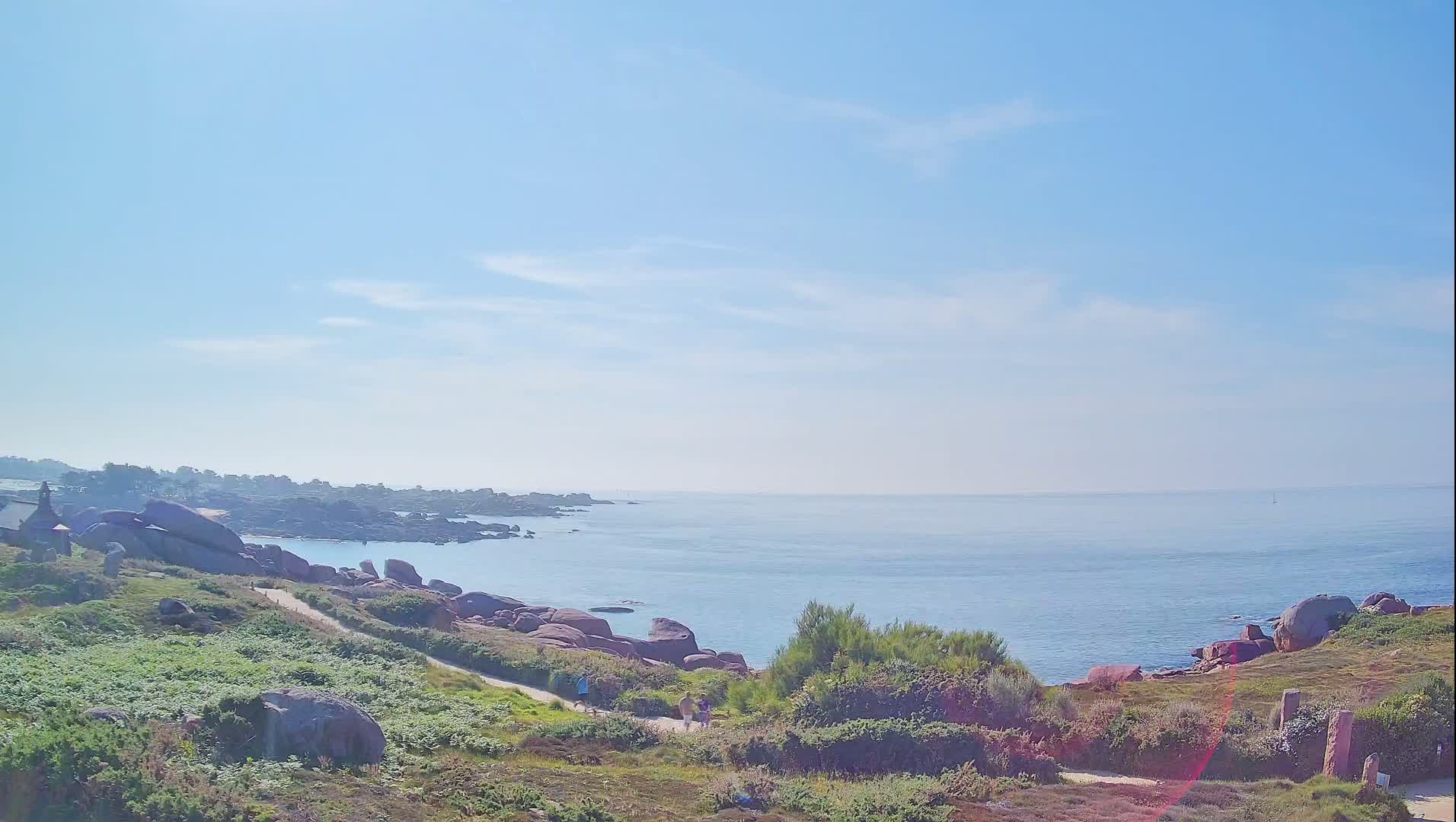A mostly sunny day reveals a rocky coastline with a calm ocean, a path winding through low-lying vegetation, and a few people strolling along the shore.