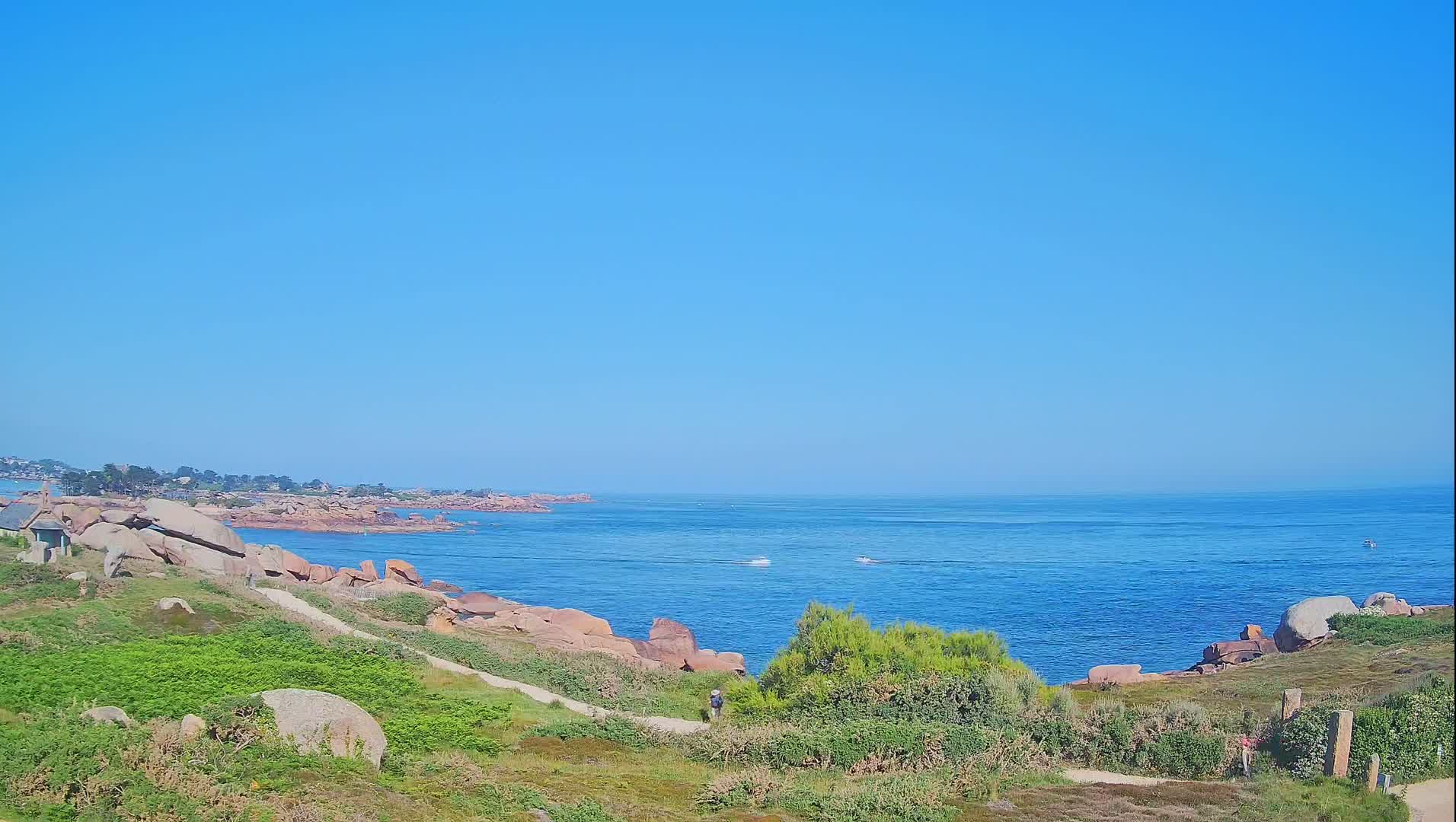 A person walks along a path overlooking a rocky coastline and calm blue ocean under a clear blue sky.