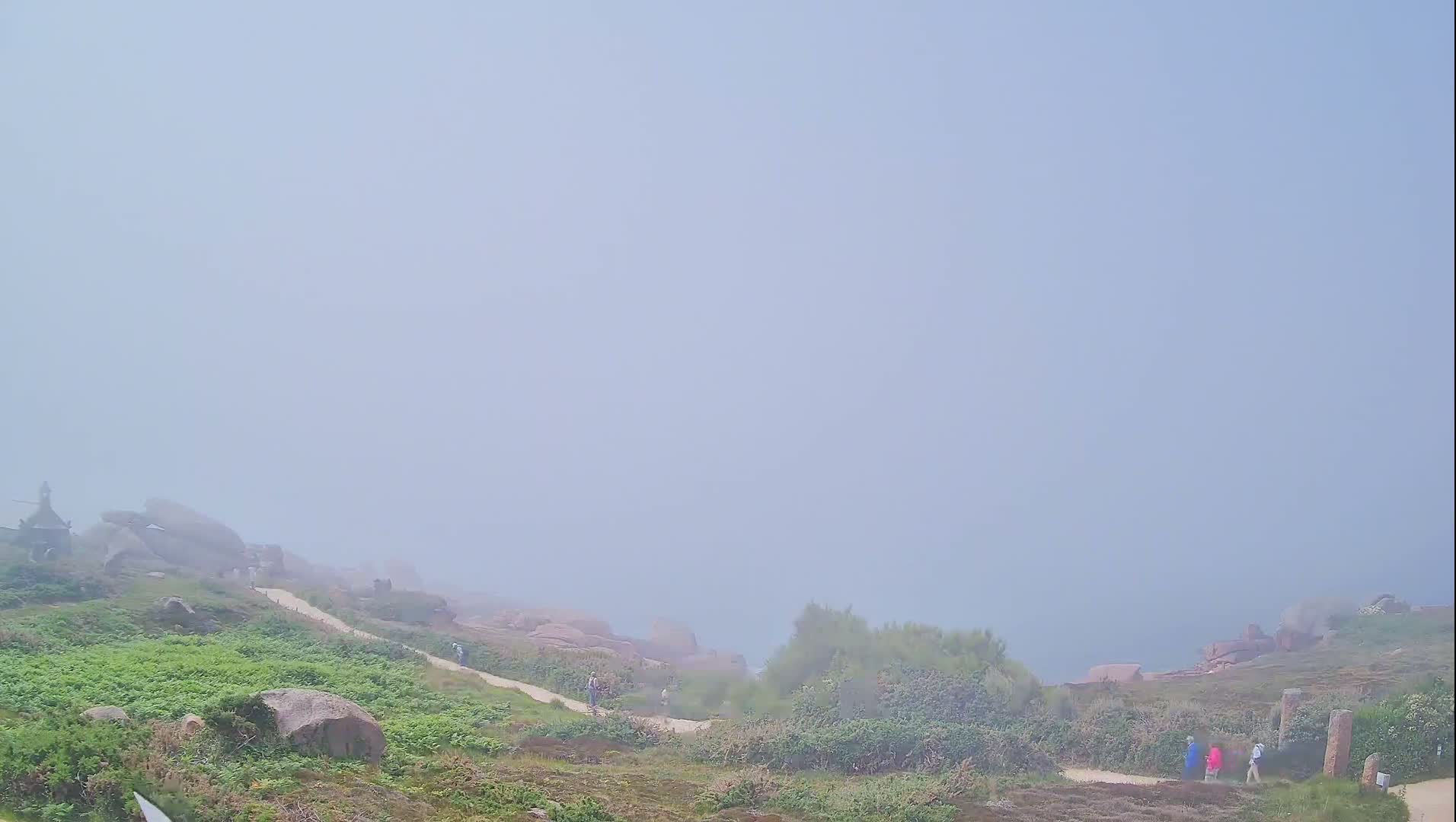 A foggy coastal scene shows a path winding through low-lying vegetation and rocky outcrops, with a few people walking along the path.