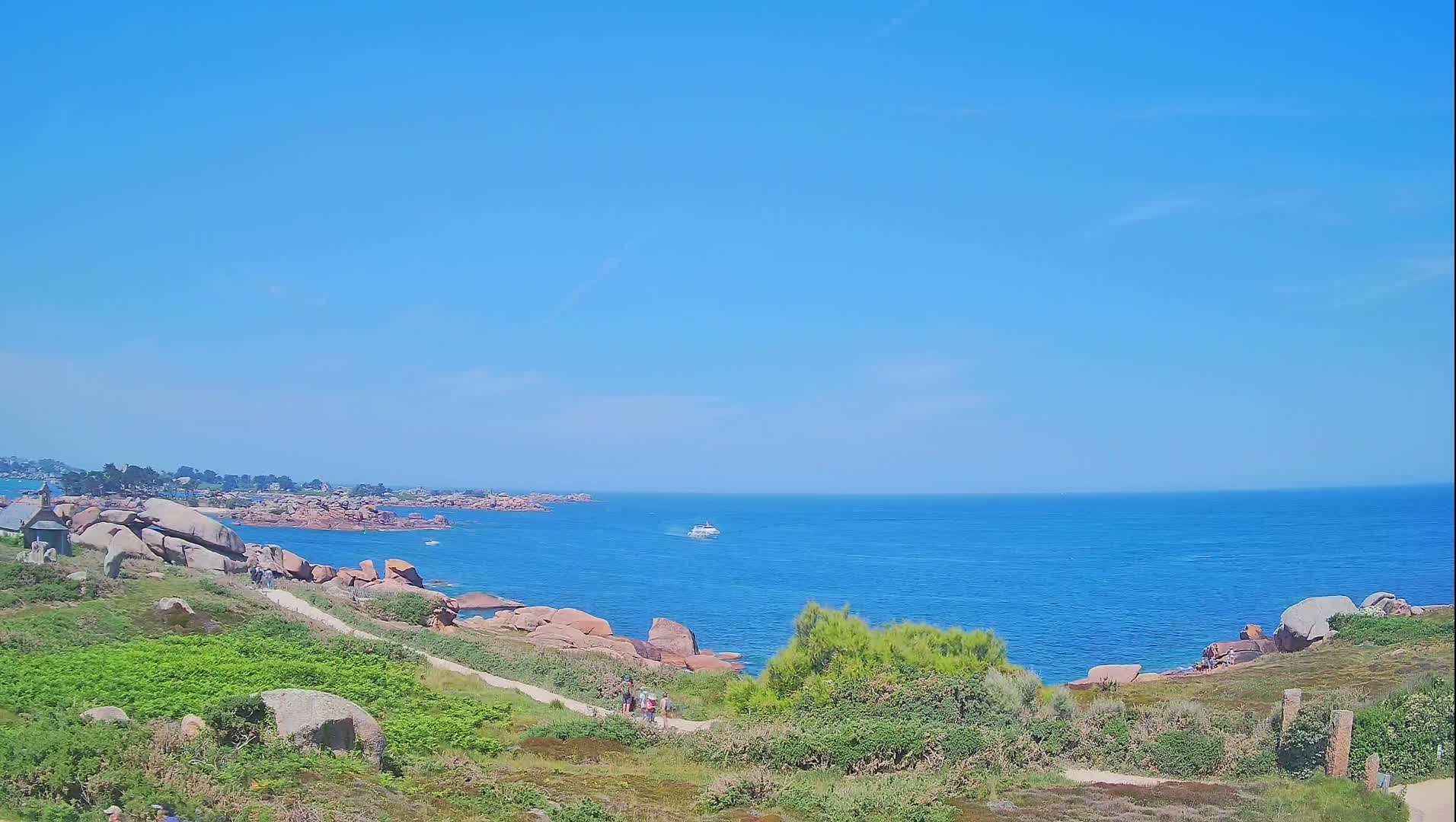A coastal path winds through green vegetation overlooking a calm blue ocean dotted with pink rocks under a clear blue sky.