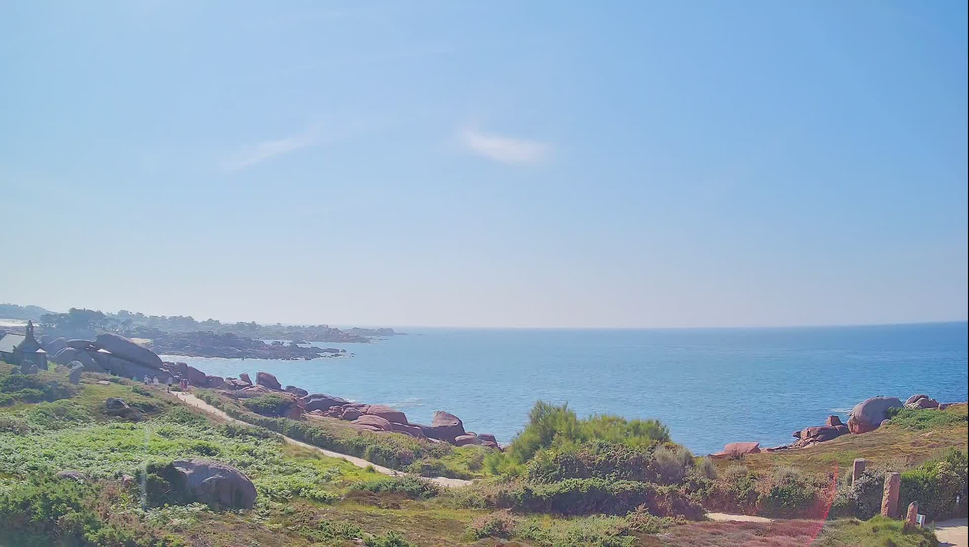 A coastal path winds through green vegetation and pink granite rocks overlooking a calm, blue ocean under a clear, sunny sky.