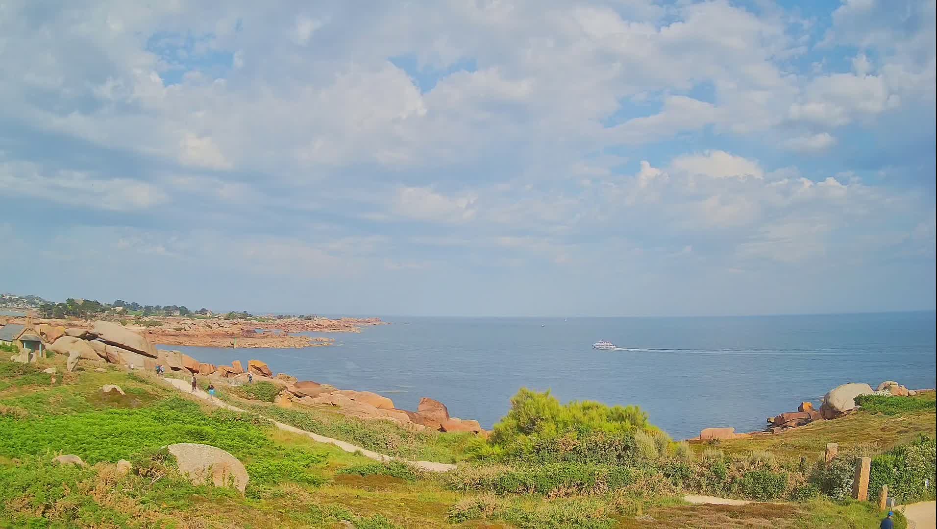 A partly cloudy day reveals a coastal scene with pink rocks, green vegetation, a path with people walking, and a boat on the calm sea in the distance.