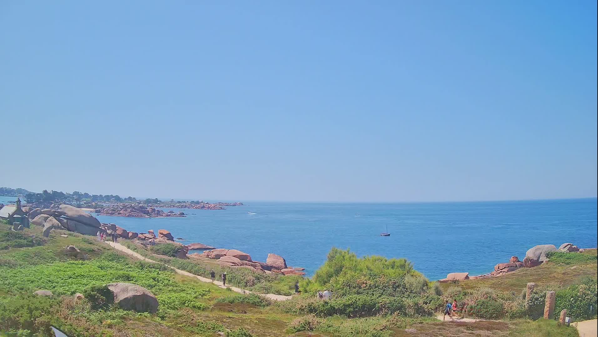 A coastal path winds through grassy hills dotted with pink granite boulders, overlooking a calm blue ocean under a clear sky.