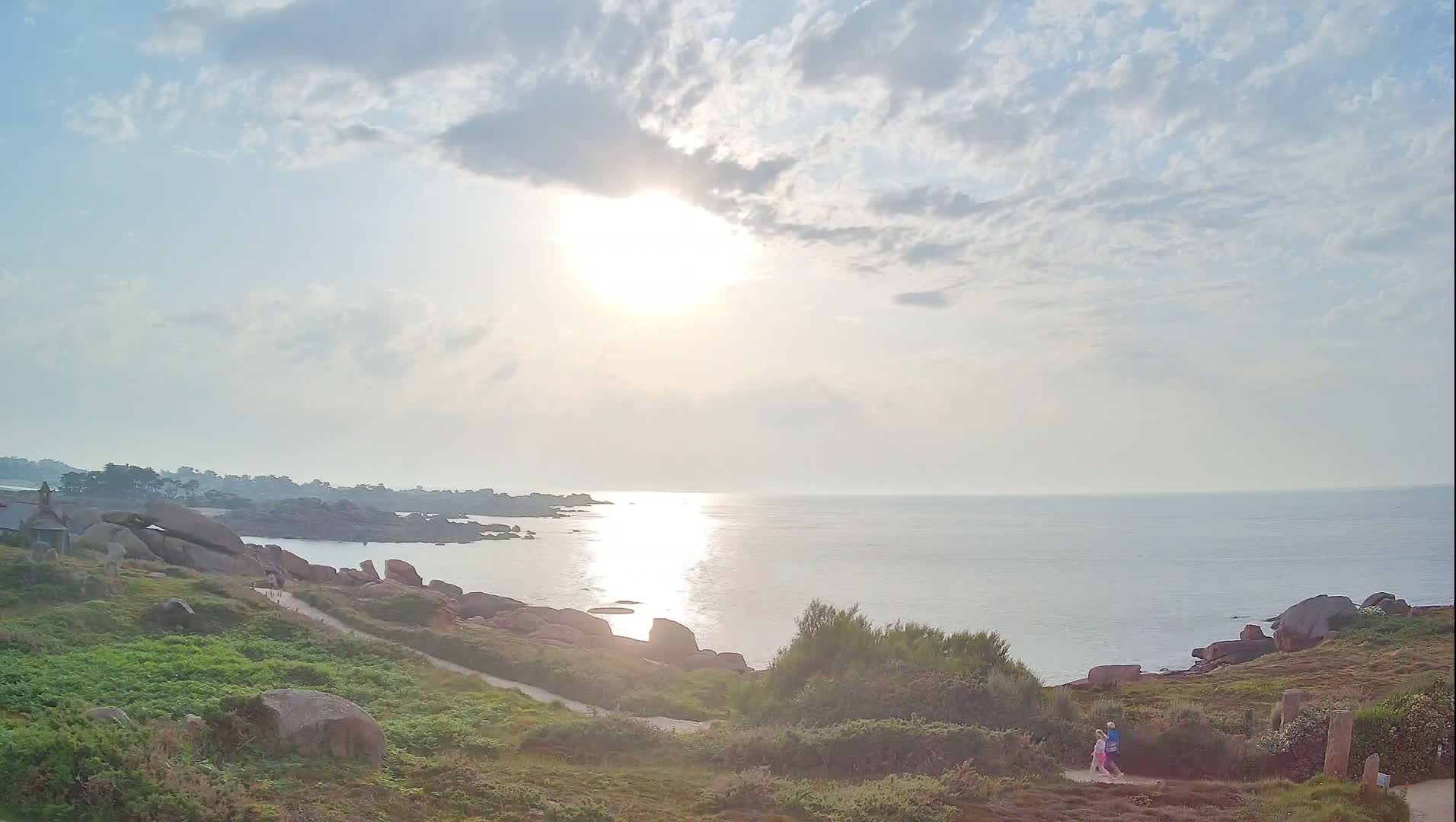 A calm, sunlit ocean scene with a rocky coastline, green vegetation, and two people walking on a path.