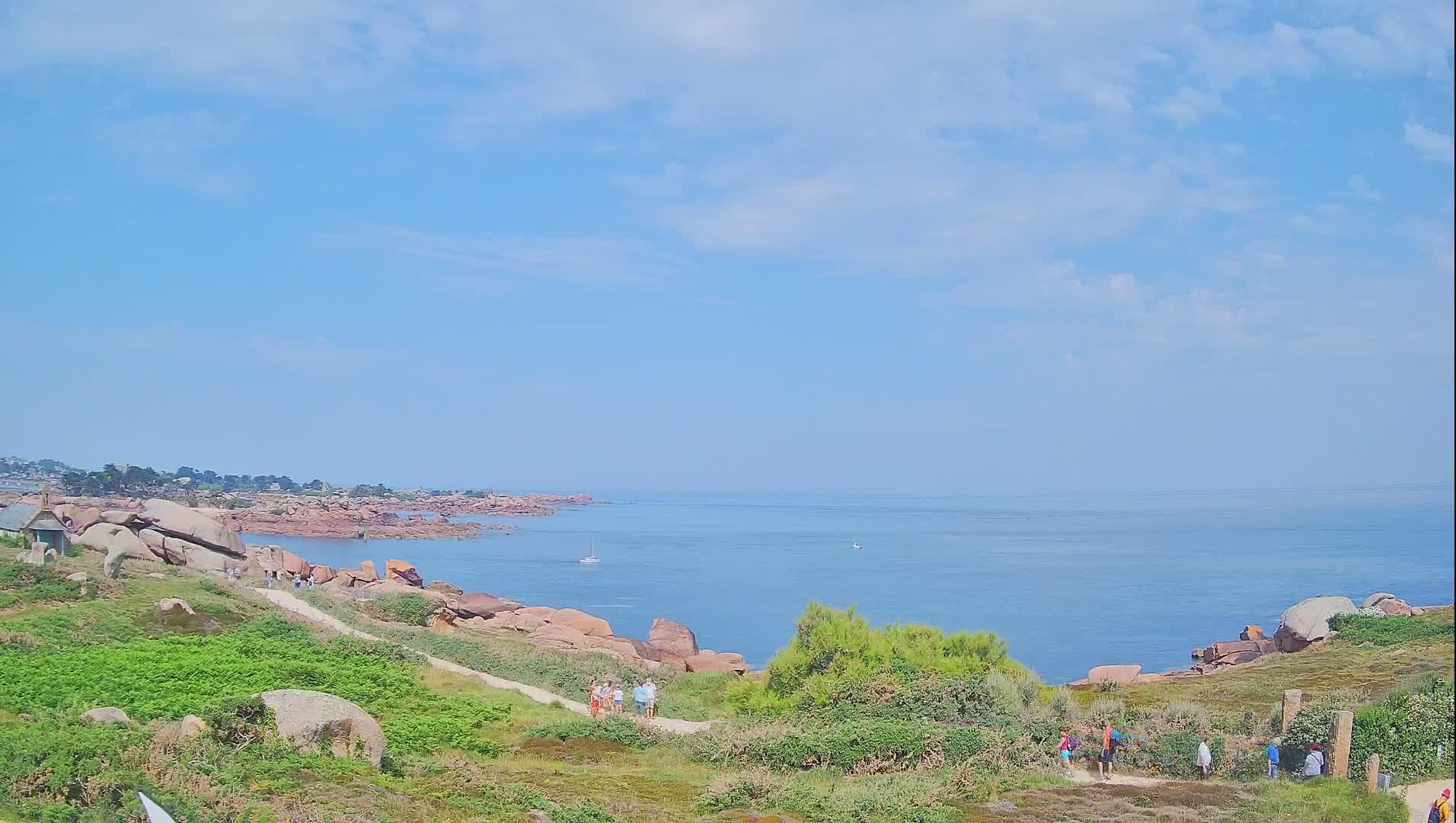 A coastal path winds through green vegetation past pink granite boulders toward a calm, blue ocean under a mostly sunny sky.