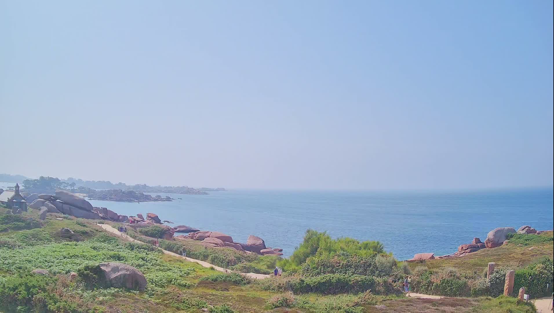A coastal path winds through green vegetation past pink granite boulders overlooking a calm blue sea on a clear, sunny day.