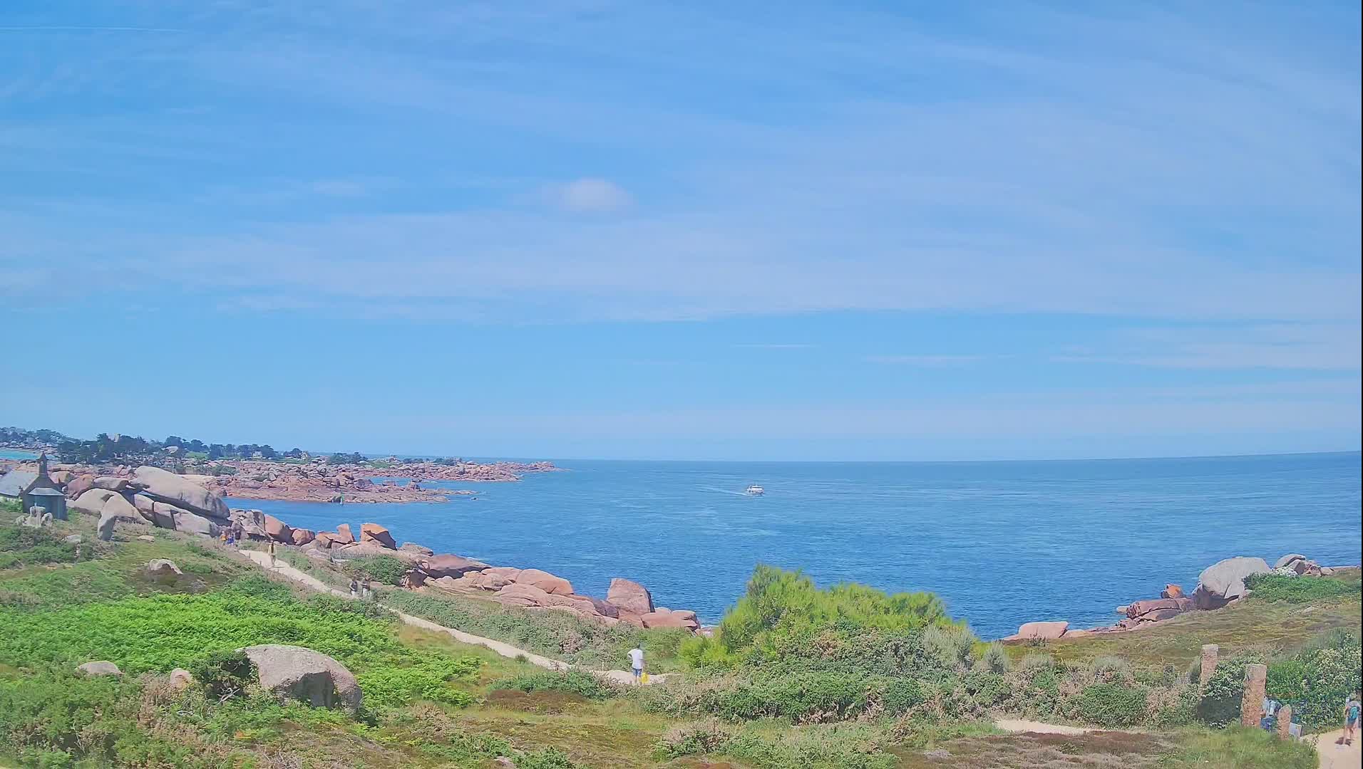 A coastal path winds through green vegetation alongside pink granite rocks and a calm blue sea under a mostly clear sky.