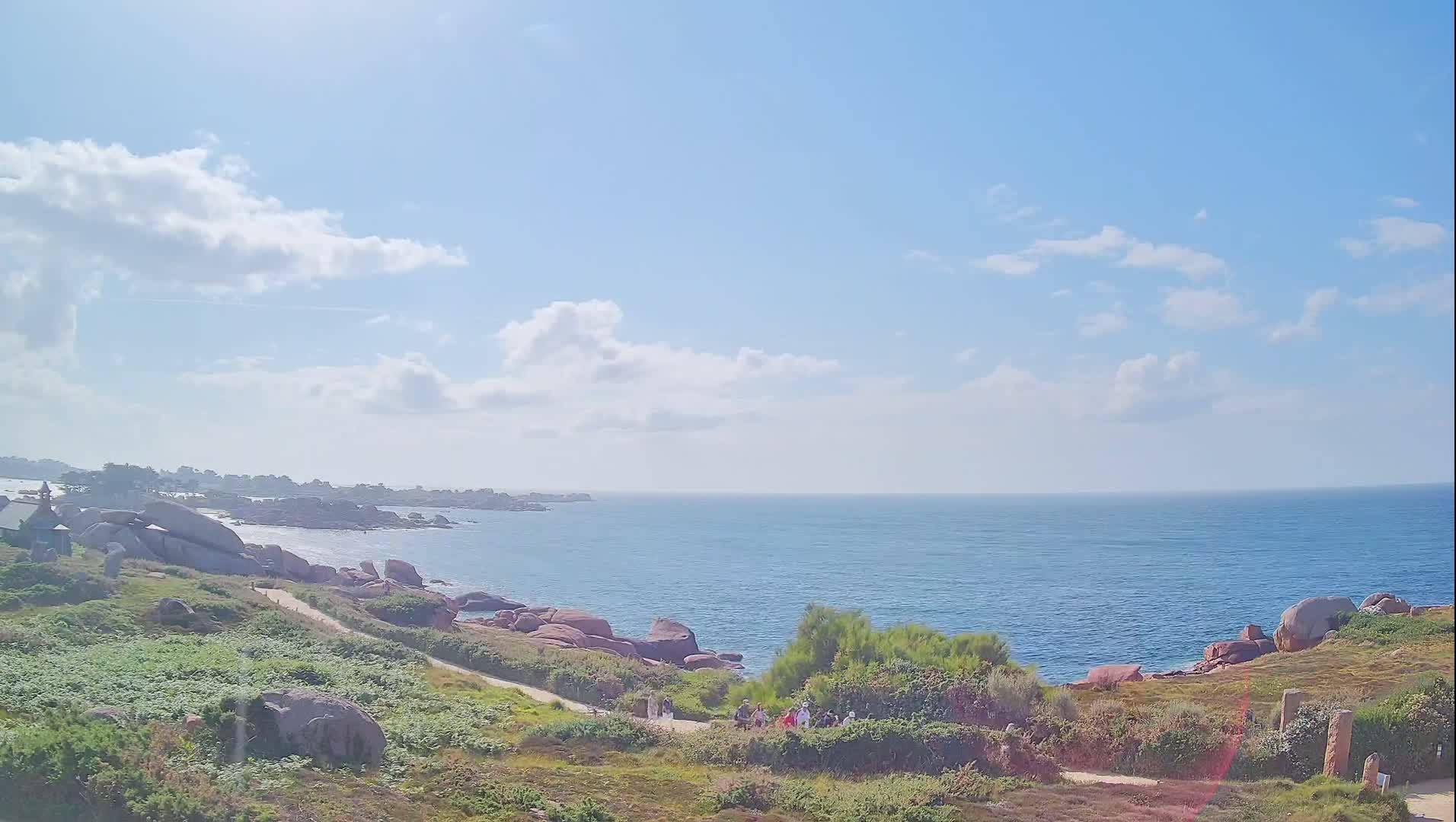 A partly sunny day reveals a coastal scene of pink granite rocks, green vegetation, a calm ocean, and a few people walking along a path.