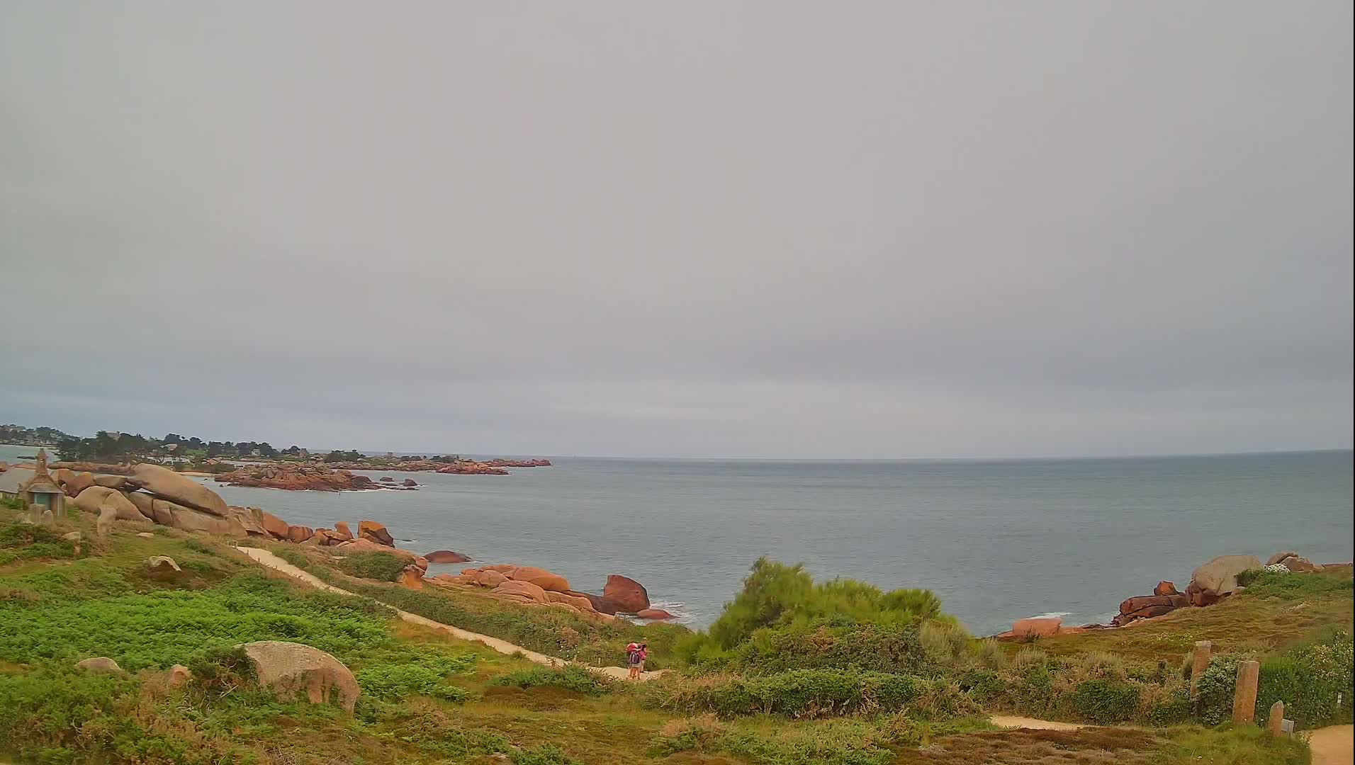 A coastal path winds through low-lying vegetation toward a calm, grey sea under an overcast sky, with pink rocks and a small settlement visible in the distance.