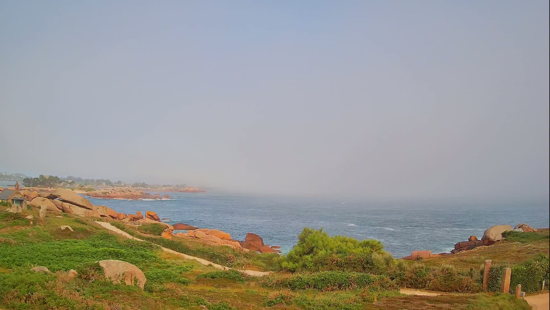 A hazy coastal scene shows a rocky shoreline with green vegetation, a path leading down to the water, and a calm sea extending to the horizon.