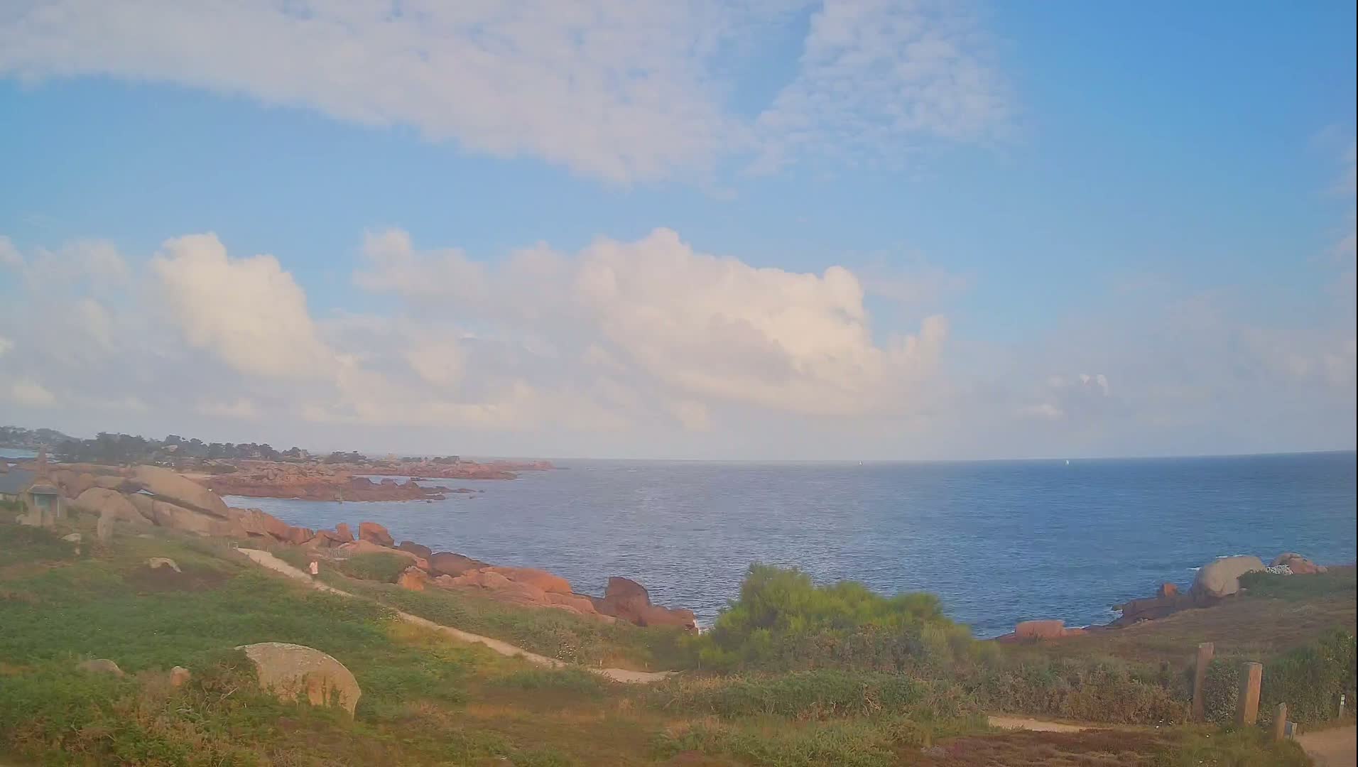 A partly cloudy sky overlooks a rocky coastline with a calm ocean and a path leading down to the water.