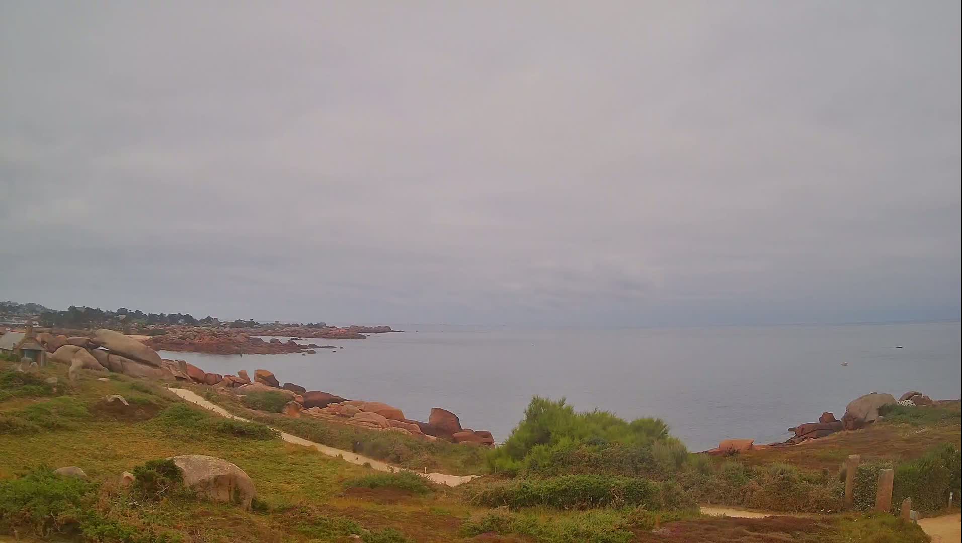 A rocky coastline with a calm, gray sea under an overcast sky is seen from a grassy hillside with a path leading down to the water's edge.