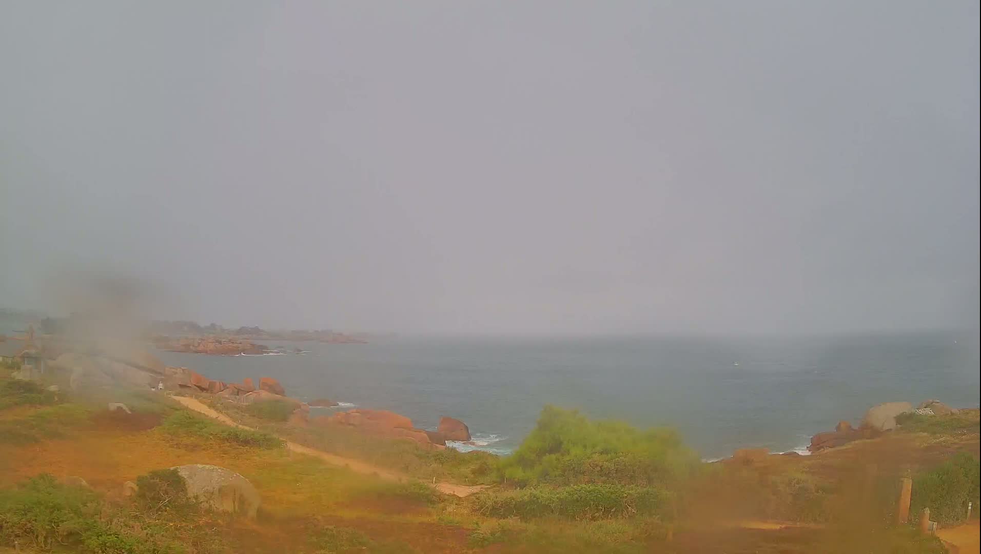 A hazy, overcast coastal scene shows a rocky shoreline with a path leading down to the sea.