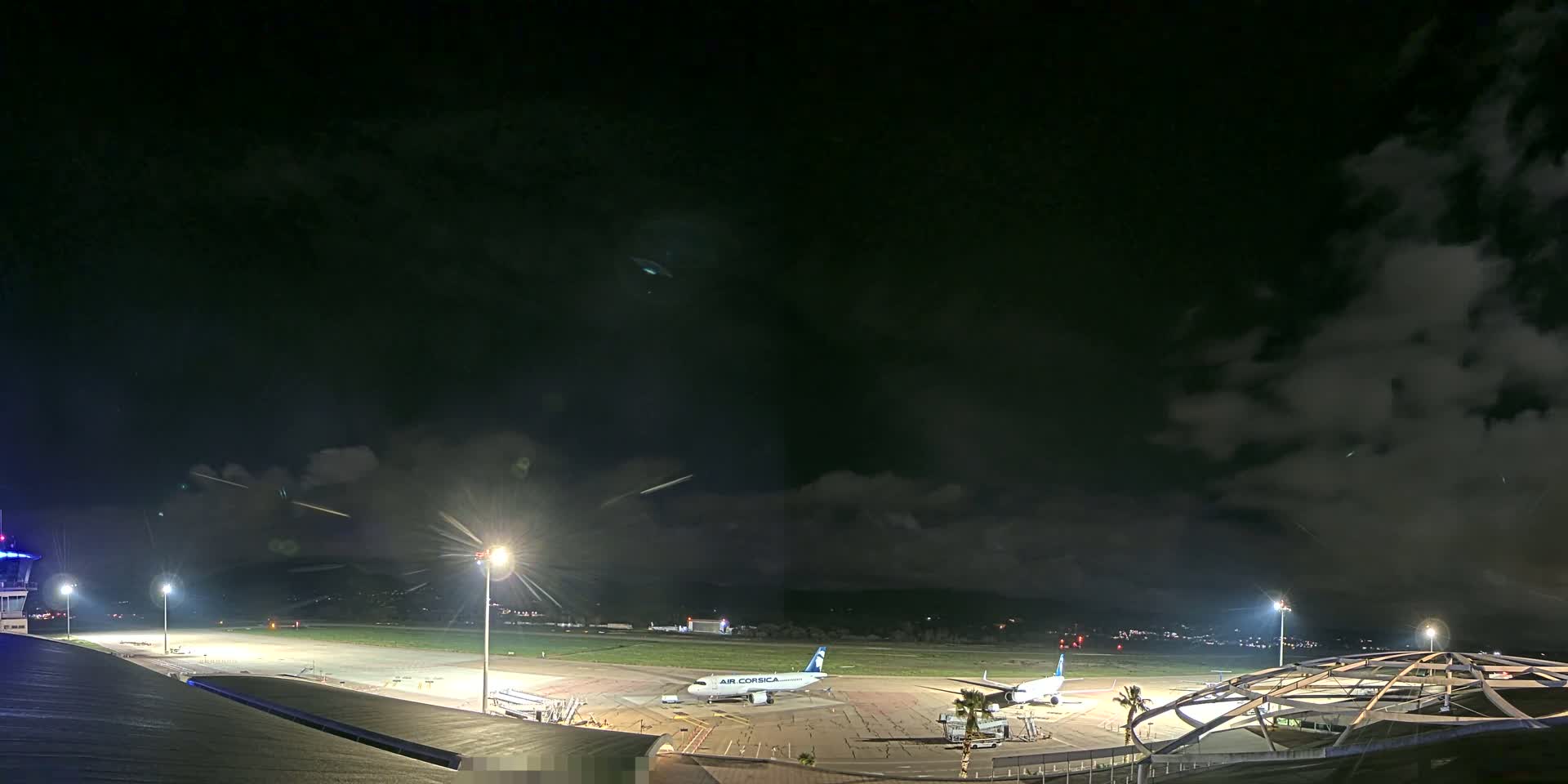 A nighttime view of an airport tarmac with two airplanes parked, under a partly cloudy sky.