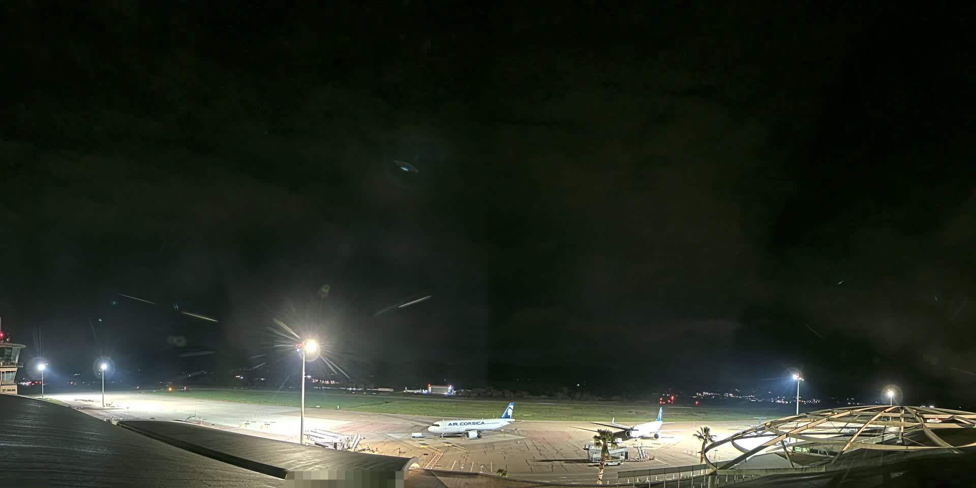 A nighttime view of an airport tarmac with two airplanes parked near a terminal building under a mostly cloudy sky.