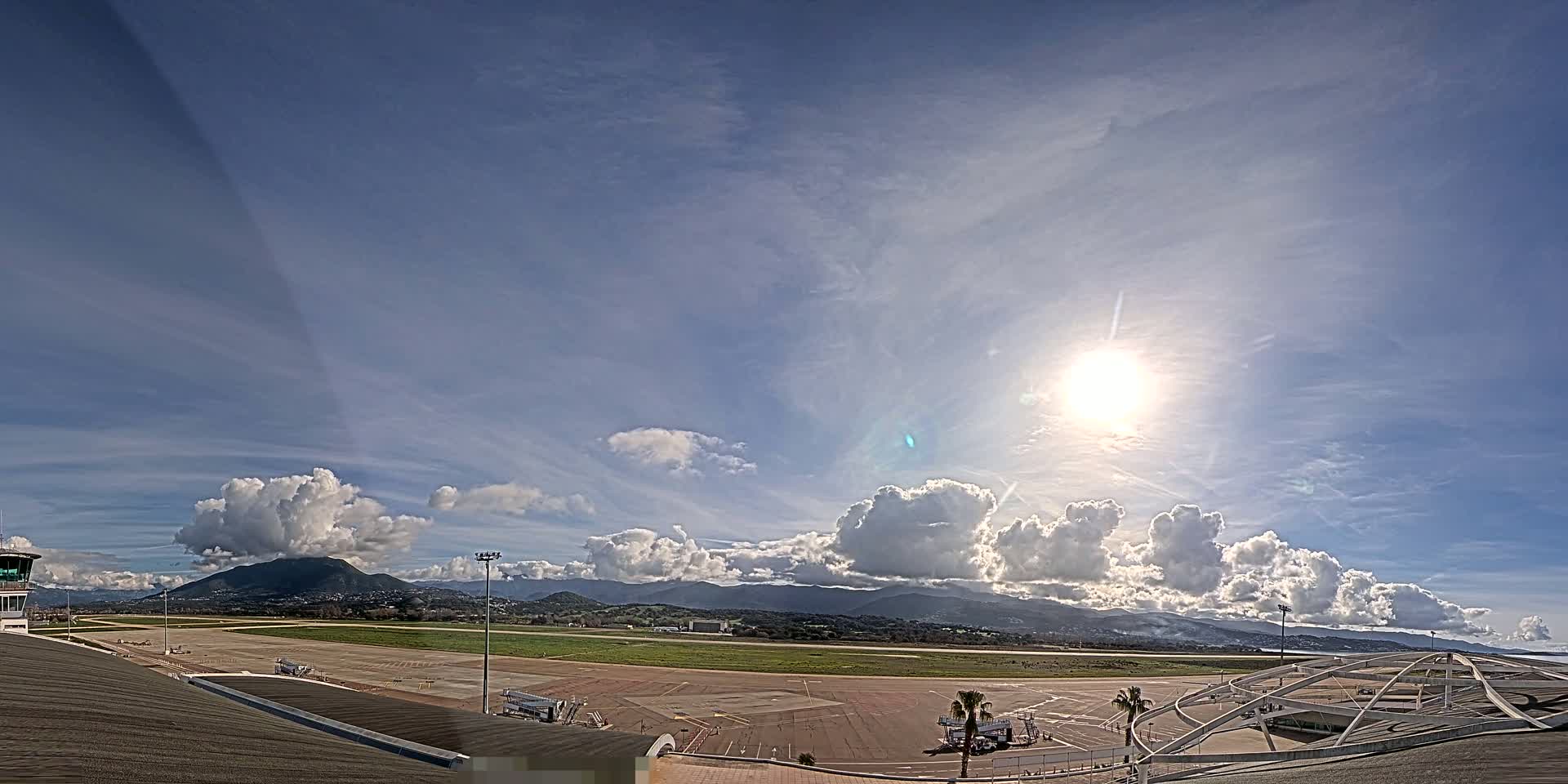 A sunny day with scattered cumulus clouds over an airport runway and surrounding landscape, including distant mountains.