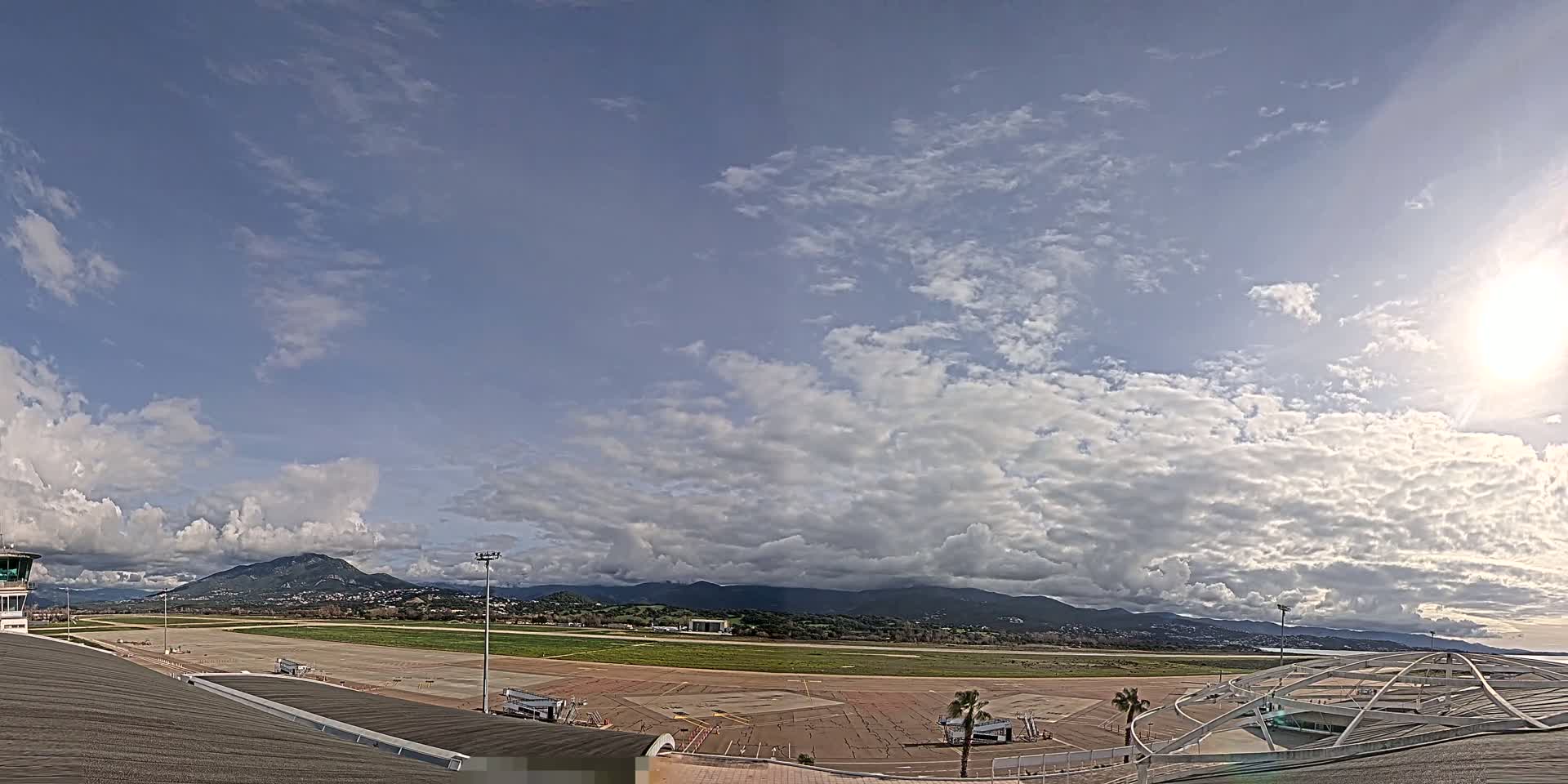 A mostly sunny day with scattered clouds over an airport runway and terminal building, with mountains visible in the distance.