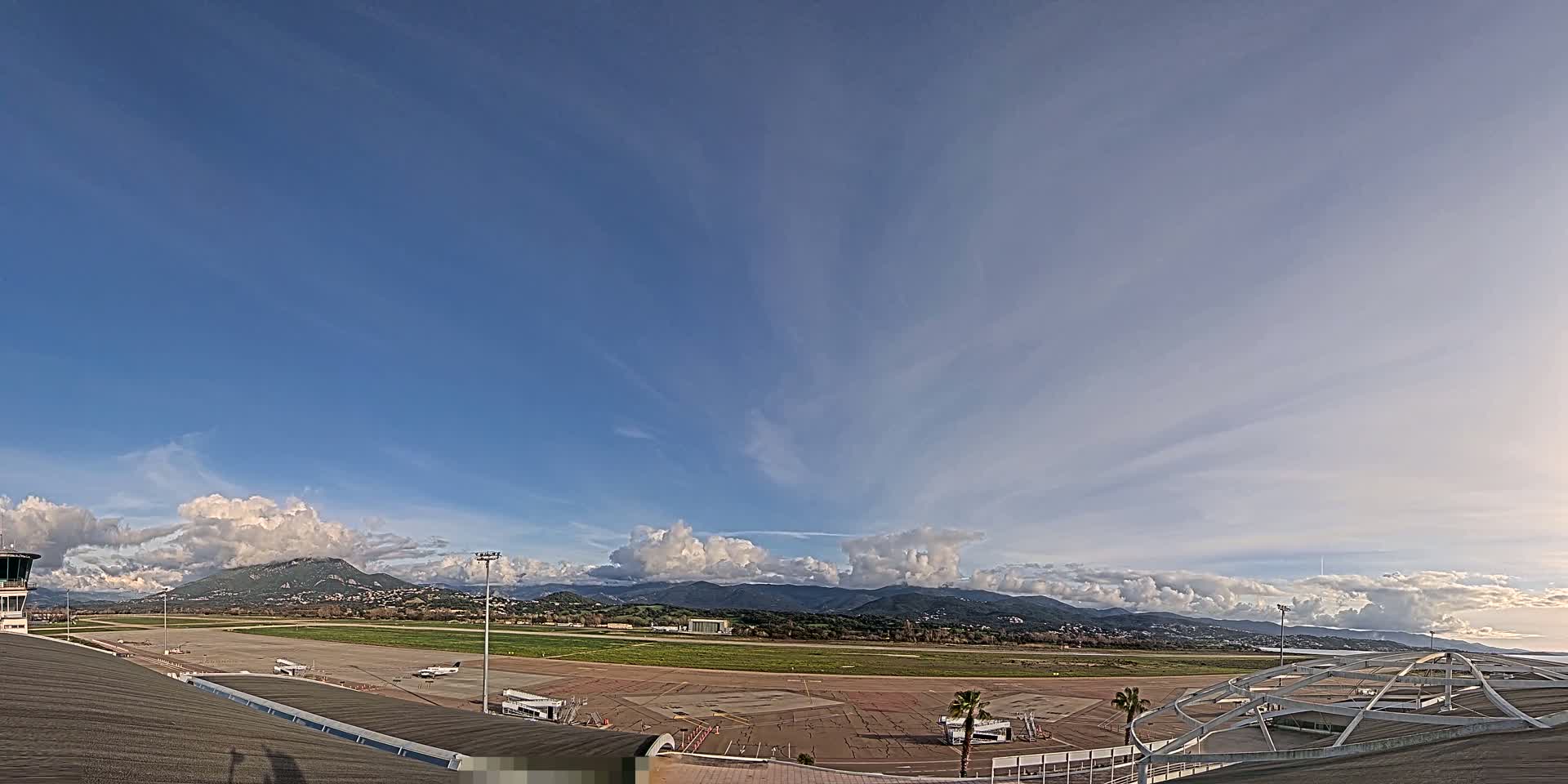 A mostly sunny day with scattered clouds over an airport runway, with mountains visible in the distance.
