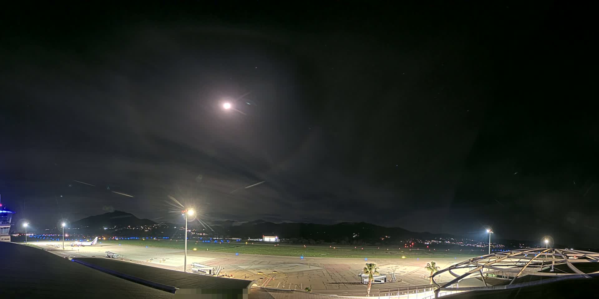 A nighttime view of an airport tarmac with a plane and several buildings under a mostly clear, moonlit sky.