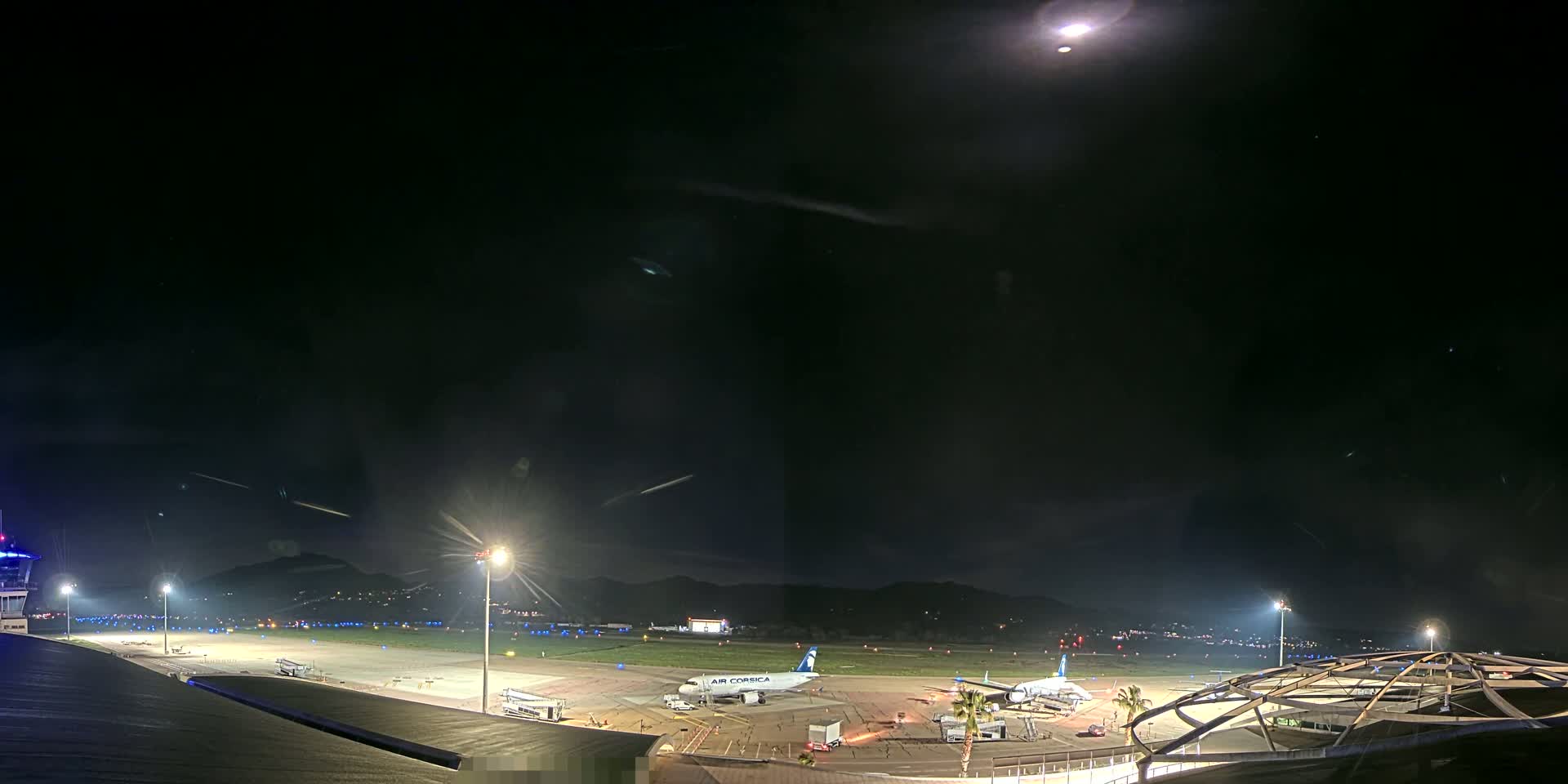 A nighttime view of an airport tarmac with a passenger plane and various ground support vehicles under a clear, moonlit sky.