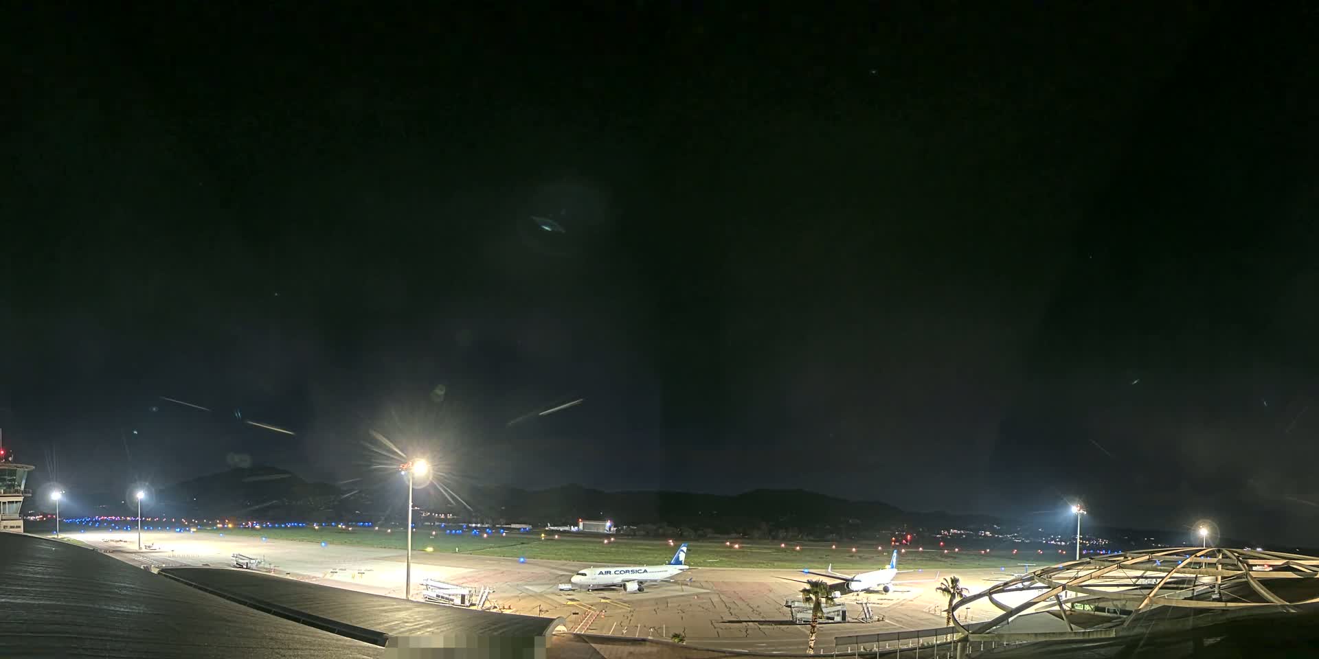 A nighttime view of an airport tarmac with two airplanes parked, illuminated by bright lights under a dark, clear sky.