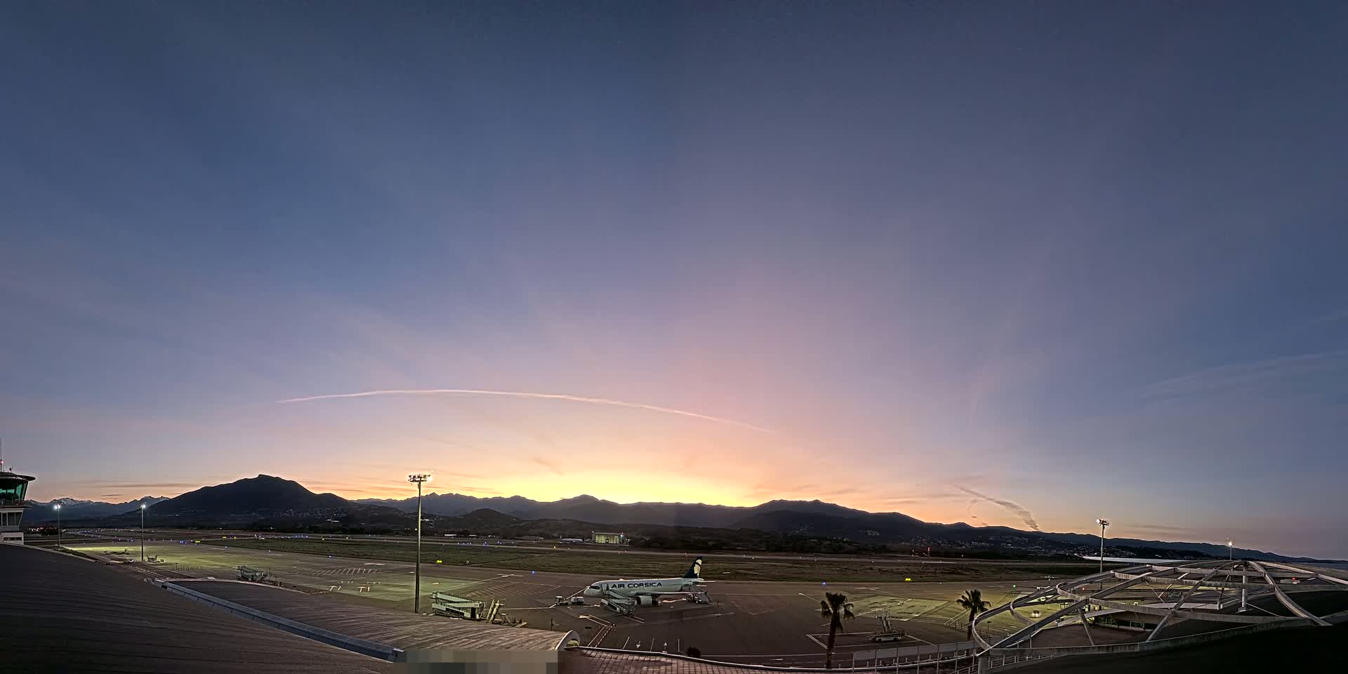 An airplane sits on an airport tarmac at sunset, with mountains visible in the distance under a clear, twilight sky.