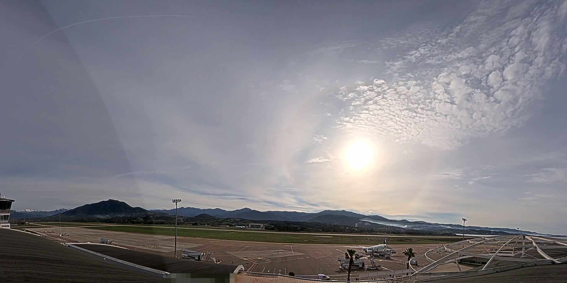 A mostly sunny day with scattered clouds over an airport runway, backed by mountains and a coastline.