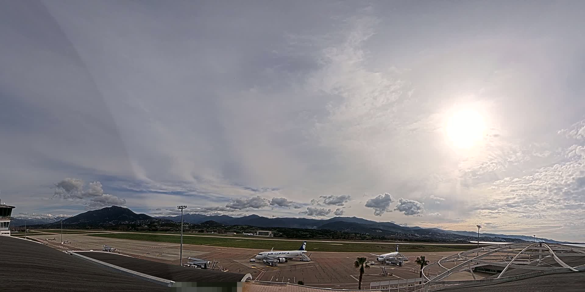 A bright, partly cloudy day at an airport shows two airplanes parked on the tarmac, a control tower, and mountains in the distance.