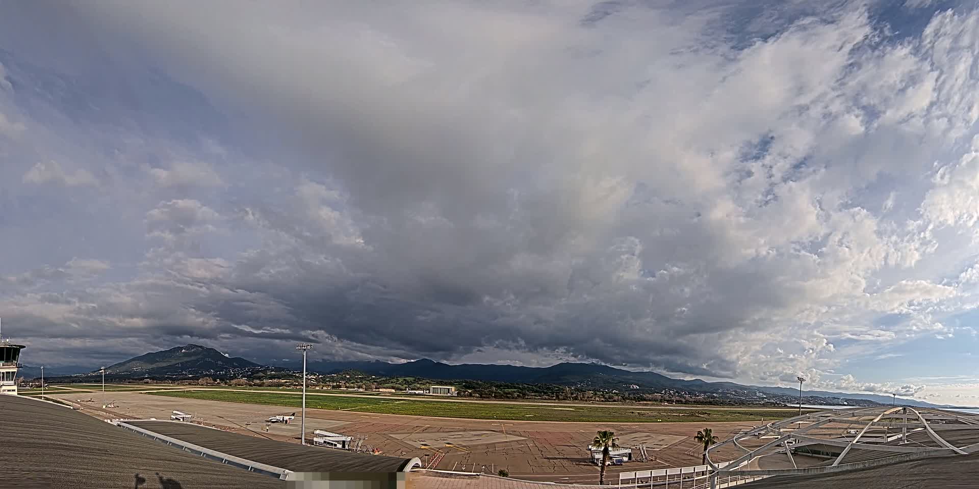 An airport tarmac with several planes, under a mostly cloudy sky with dark clouds gathering over distant mountains.