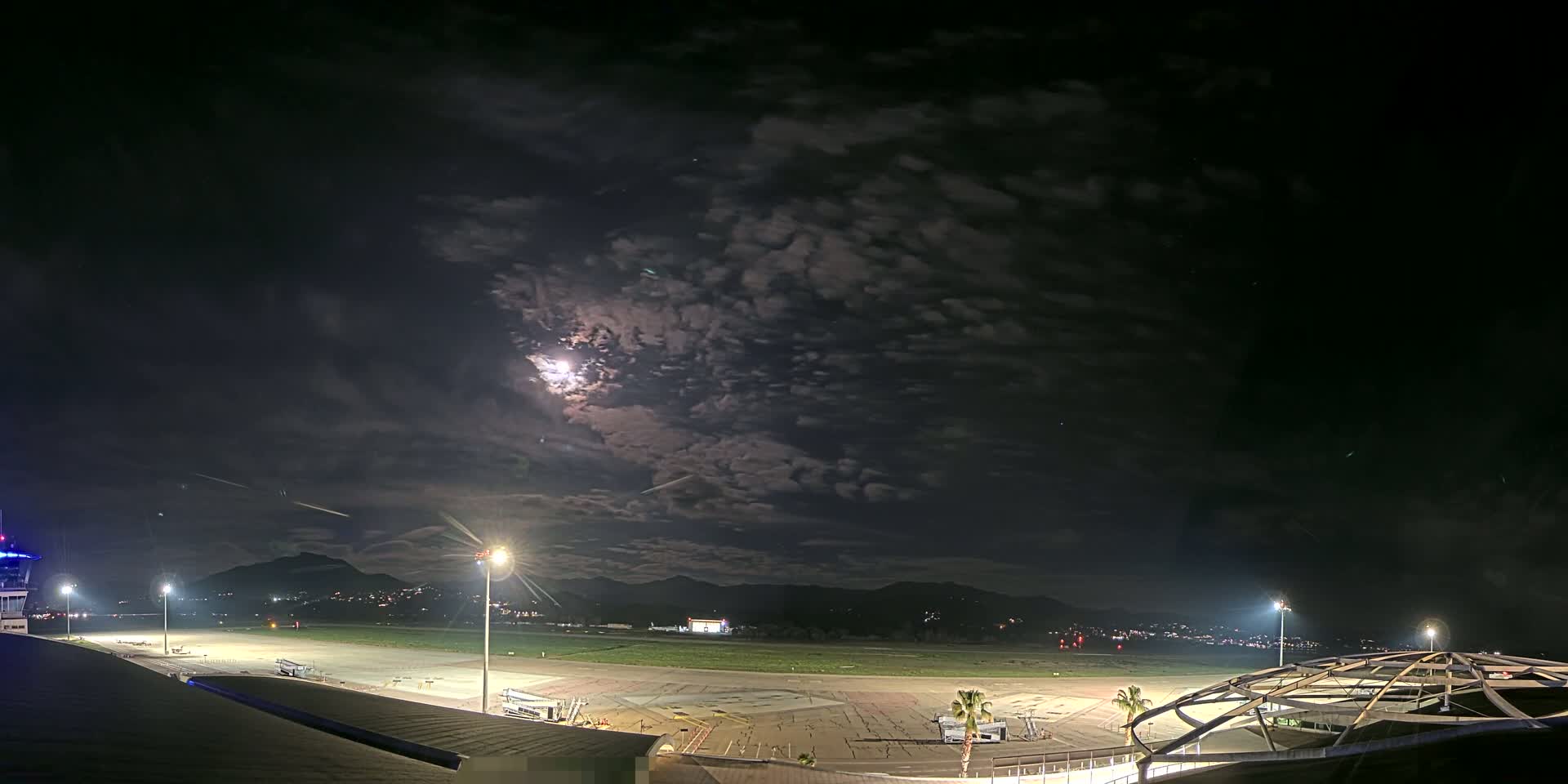 A nighttime view of a mostly empty airport runway, under a partly cloudy sky illuminated by a bright moon and airport lights, with mountains visible in the distance.