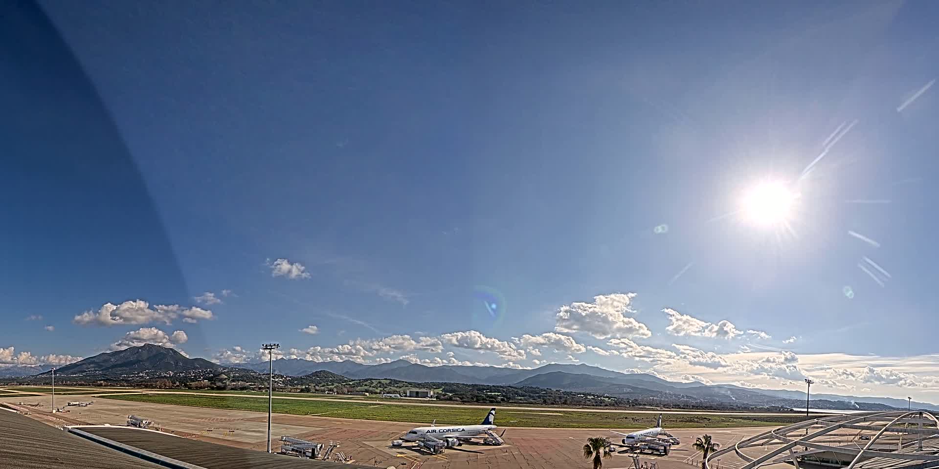A sunny day at an airport shows several airplanes parked on the tarmac with mountains in the distance.