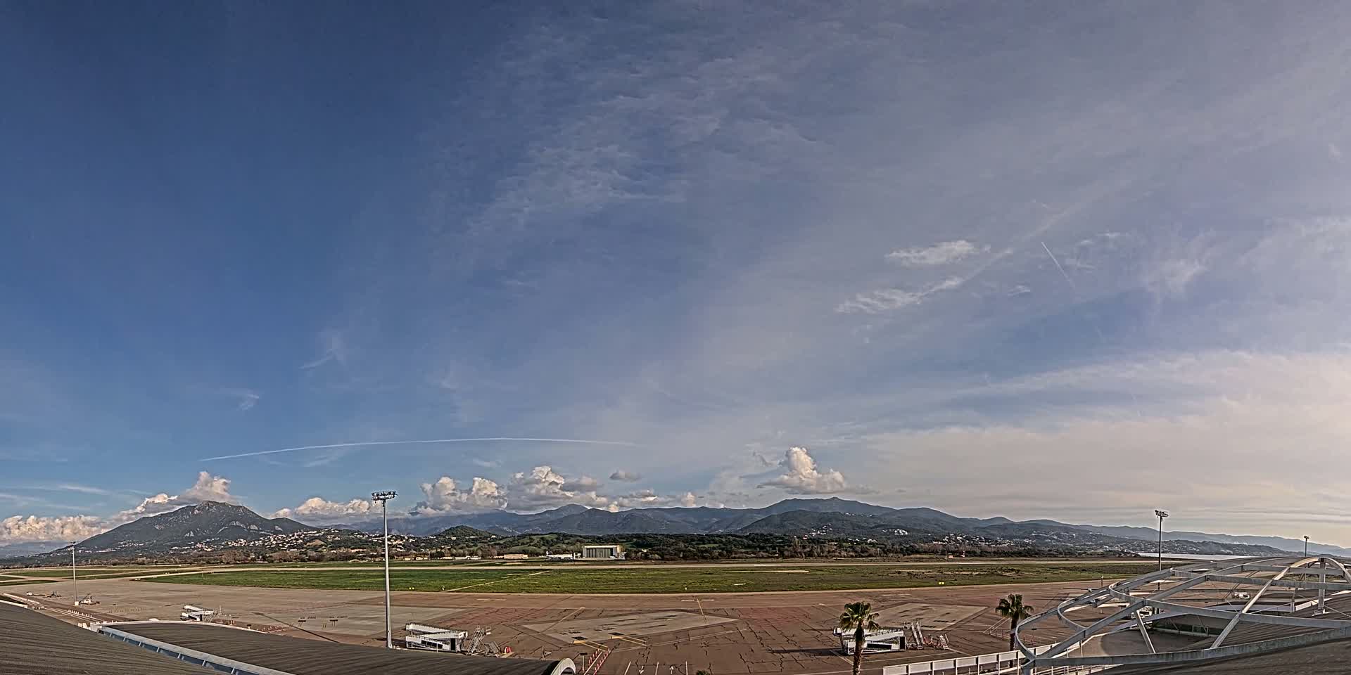 An airport tarmac with a few airplanes and jet bridges is visible beneath a mostly clear, partly cloudy sky, with mountains in the distance.