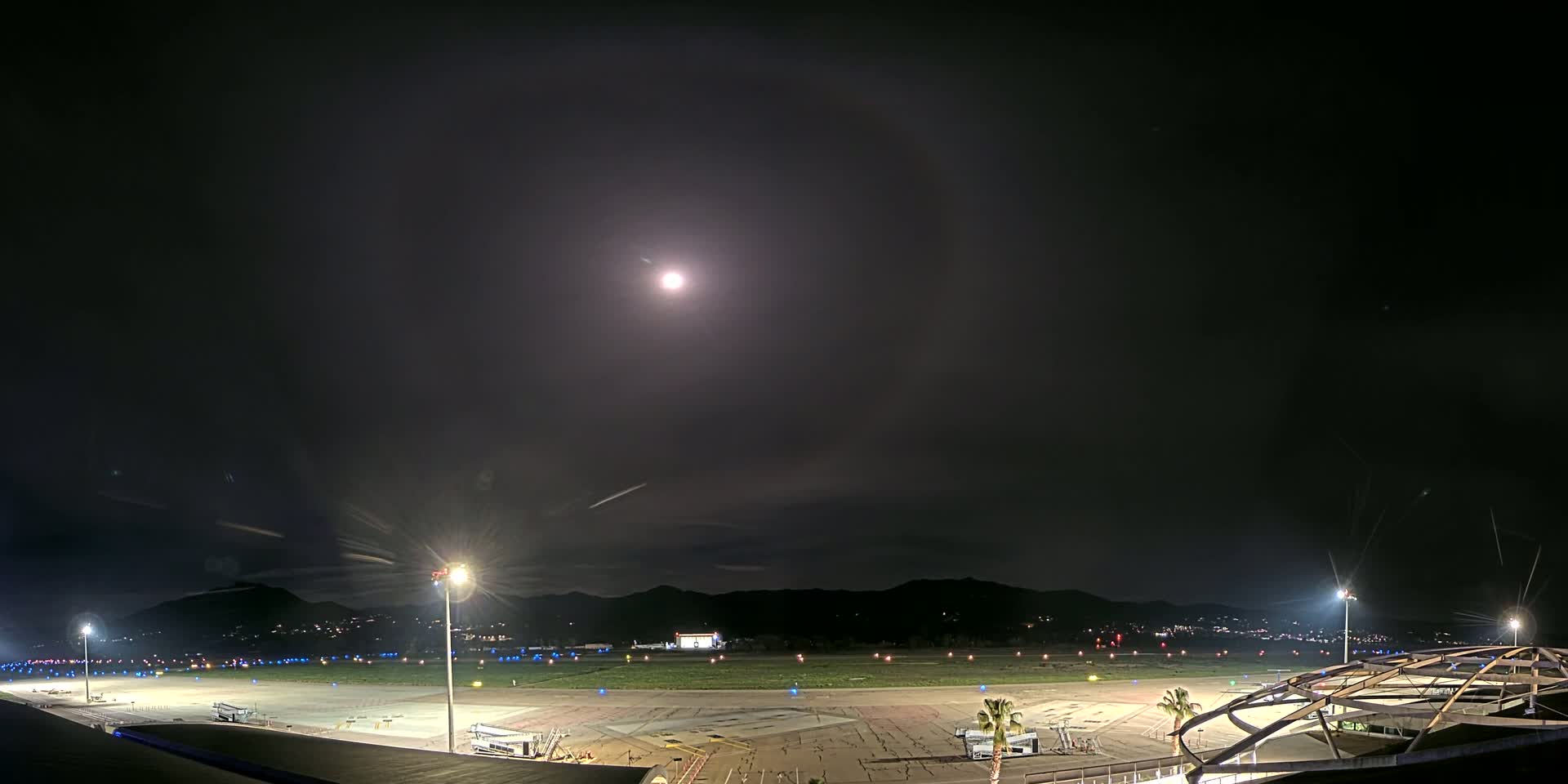 A brightly lit airport tarmac at night, under a mostly clear sky with a bright moon and faint clouds, is bordered by dark mountains in the distance.