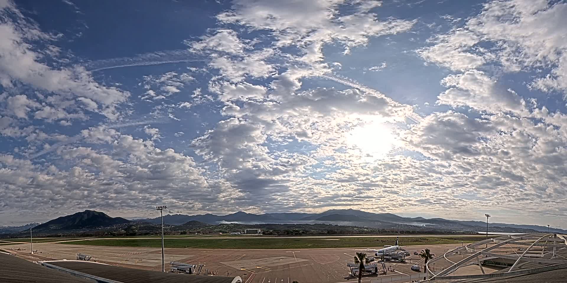 A mostly sunny day with scattered clouds over an airport tarmac, featuring a plane and distant mountains.