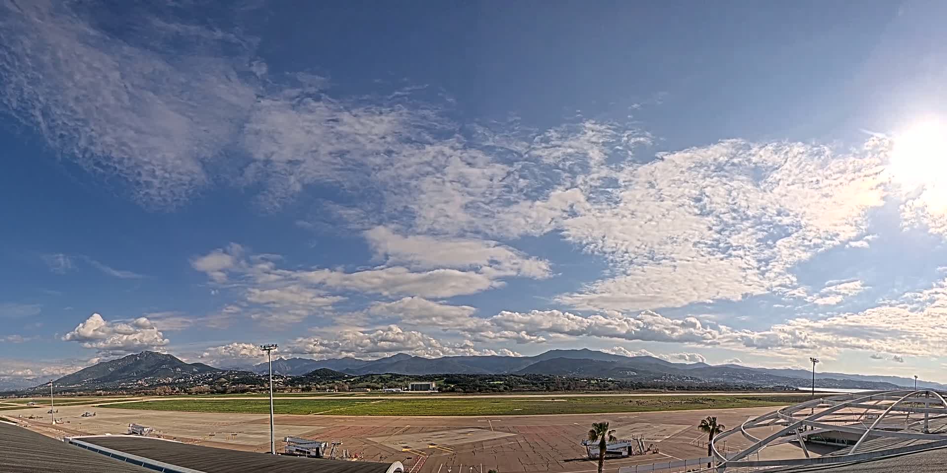 A mostly sunny day with scattered clouds over an airport tarmac and runway, with mountains visible in the distance.