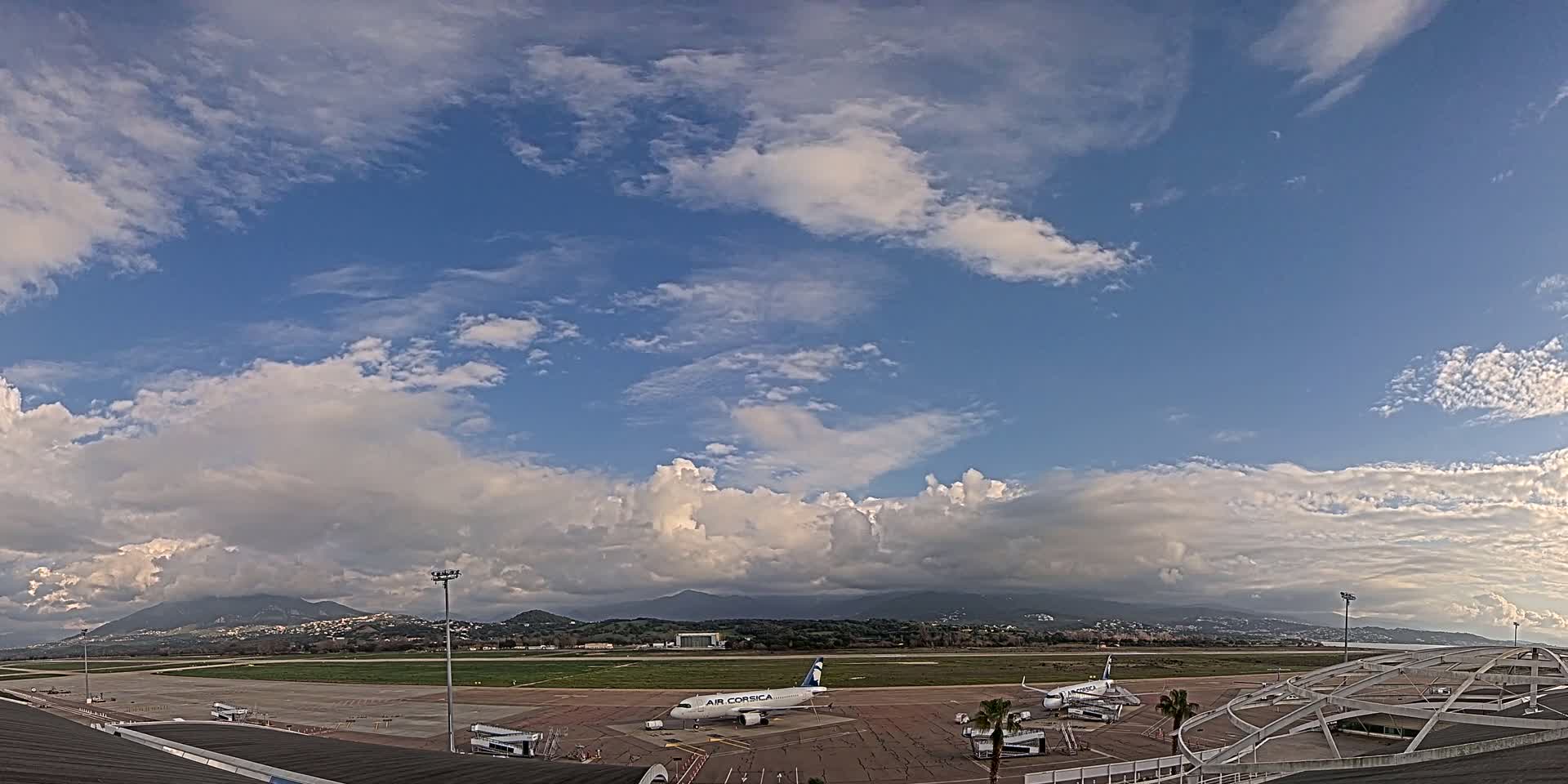 A partly cloudy sky overlooks an airport tarmac with several airplanes, including one Air Corsica plane, and mountains in the distance.