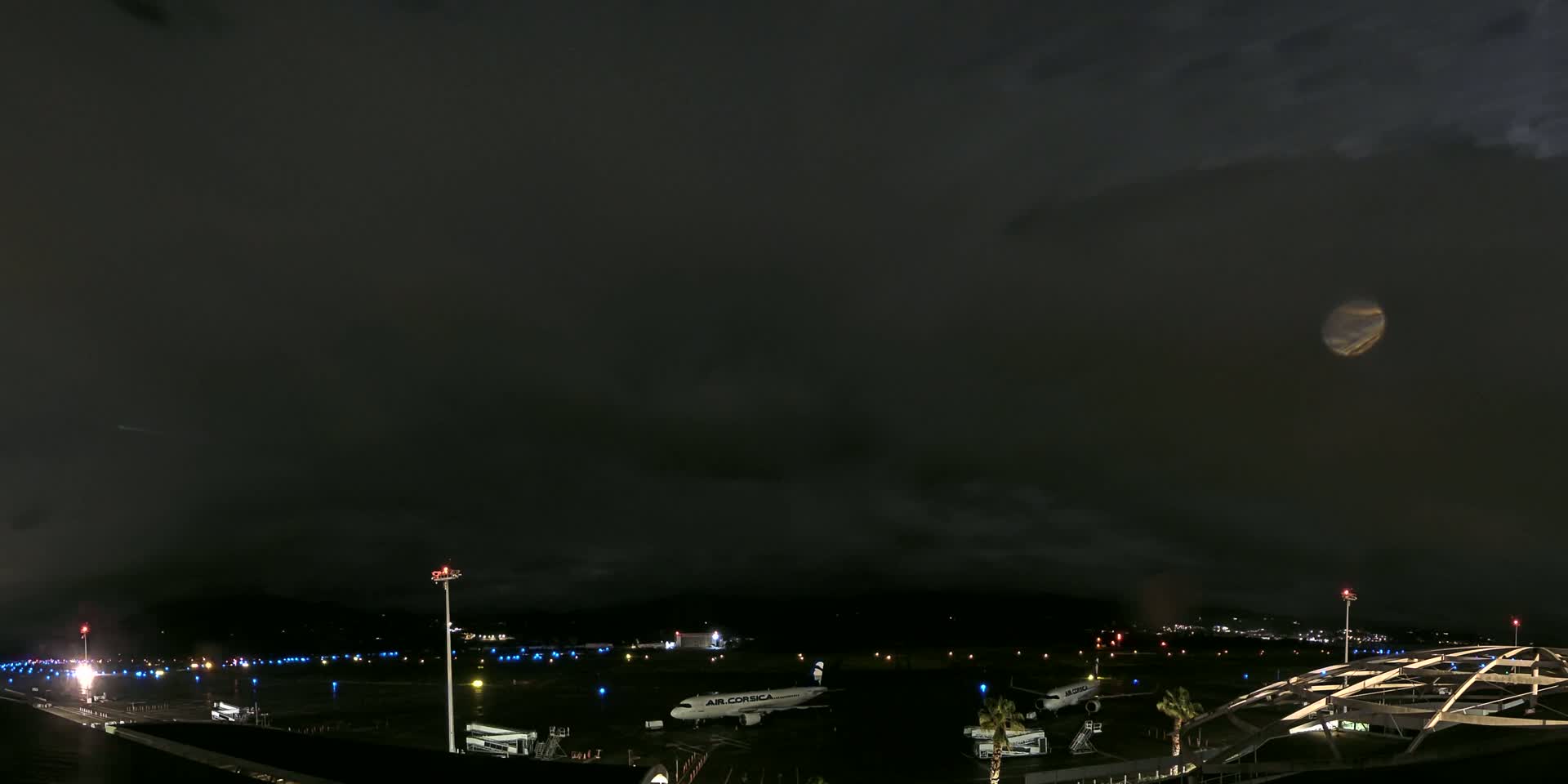 Under a dark, cloudy night sky illuminated by a bright moon, several airplanes are parked on a dimly lit airport tarmac.